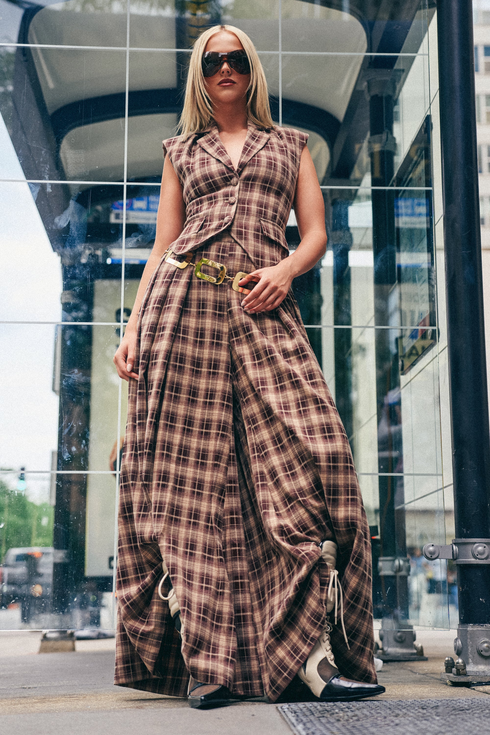 Wearing the PLAID PRINCESS TROUSER, a woman in sunglasses stands confidently at a glass-covered city bus stop. Her sleeveless, brown plaid wide-leg jumpsuit with floor-length legs embodies effortless fall streetwear style.