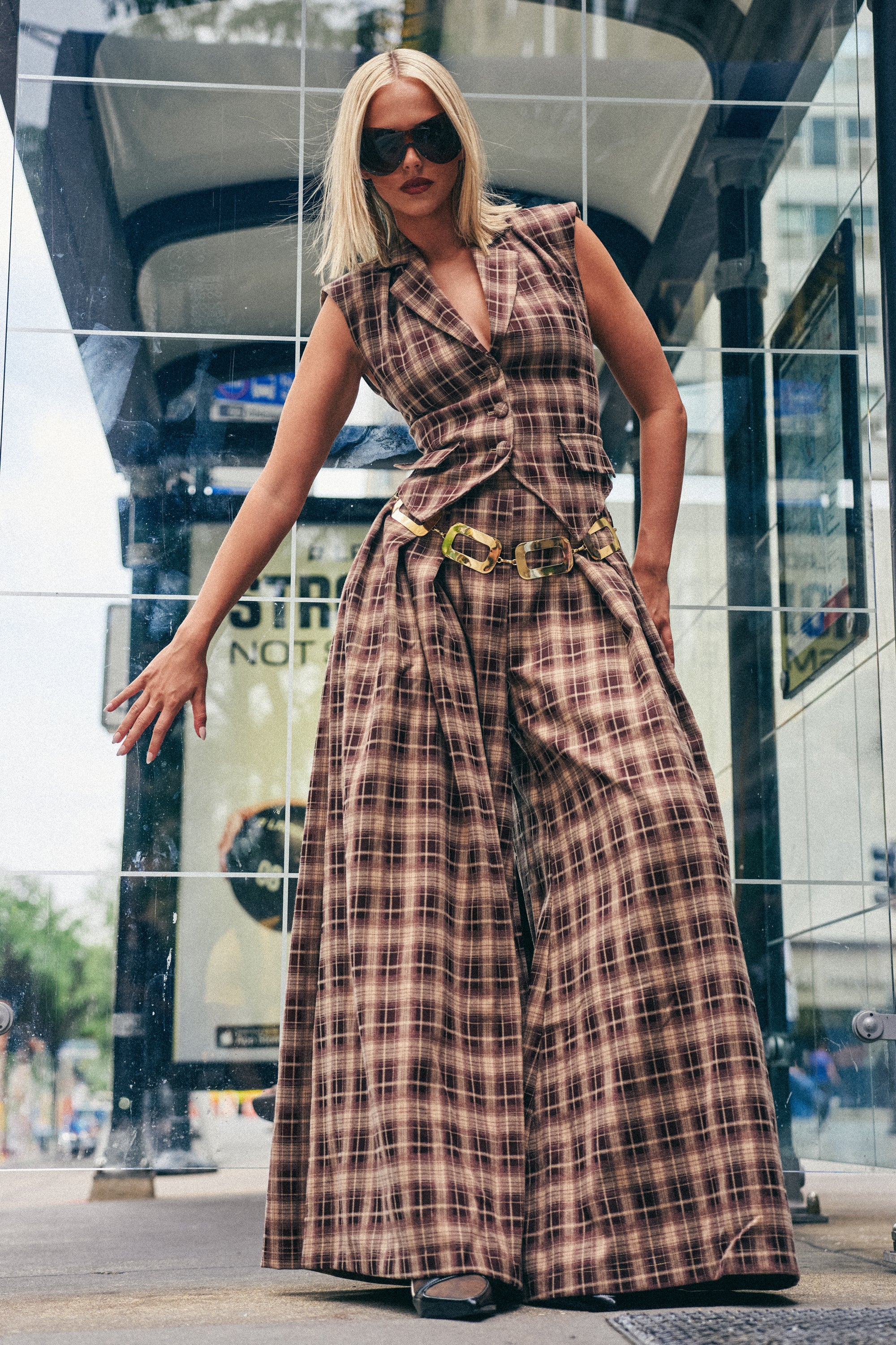 A person wearing sunglasses and the PLAID PRINCESS TROUSER, styled to capture the look of classic brown plaid wide leg trousers, stands confidently at a glass bus stop, striking a dynamic pose—a perfect example of chic fall streetwear.