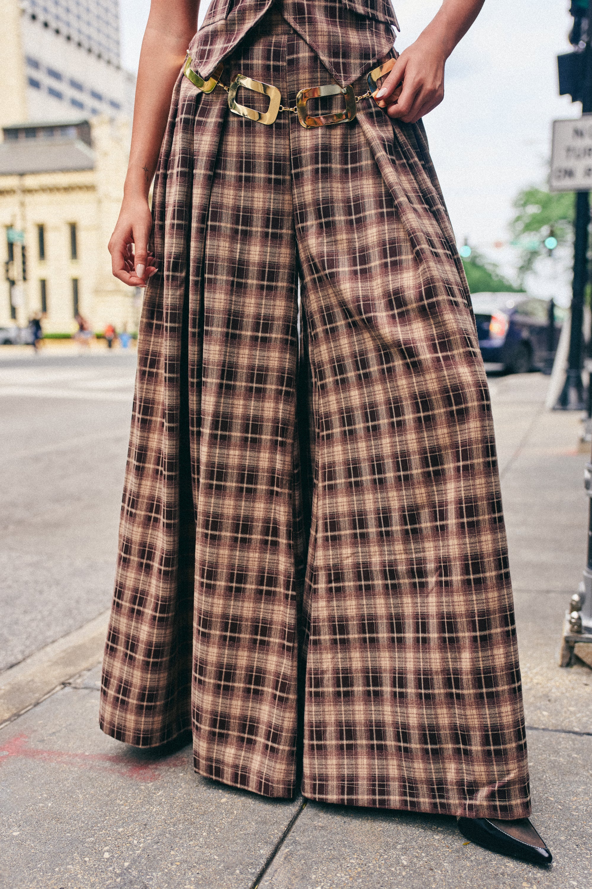 Standing on a city sidewalk, the person showcases the PLAID PRINCESS TROUSER—a high-waisted, brown plaid wide-leg pant paired with a matching top and finished with a chunky belt. Perfect for fall streetwear, the individual’s head is not visible in the image.