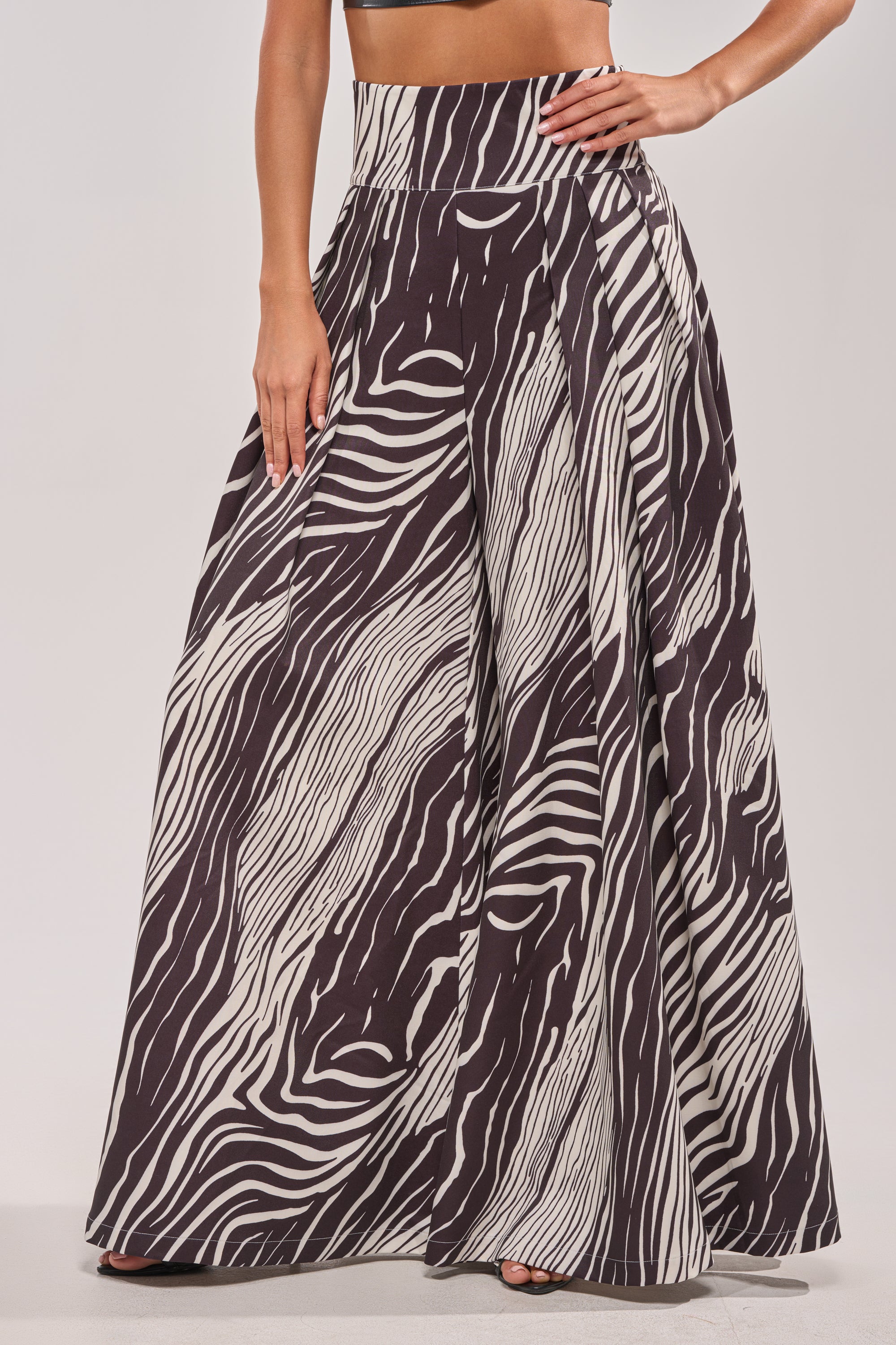 A person wears the WILD AND FREE ZEBRA TROUSER, featuring a high-rise waist, bold black and white zebra print, wide-leg floor-length silhouette, paired with a cropped top that reveals the midriff.
