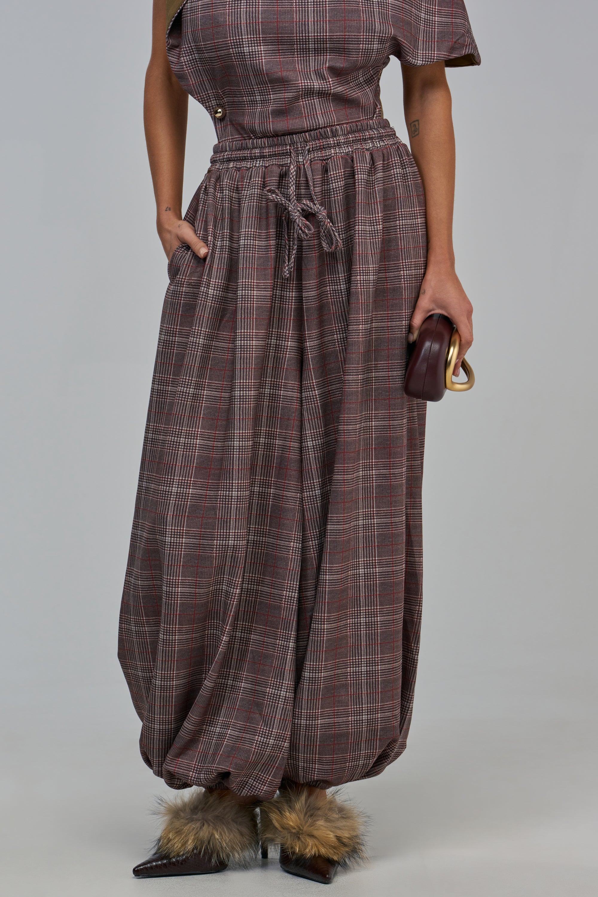 A person wears RUN THE SHOW BALLOON PANT in a high-rise, brown plaid relaxed fit with a drawstring waist and dark pointed-toe shoes with faux fur trim.