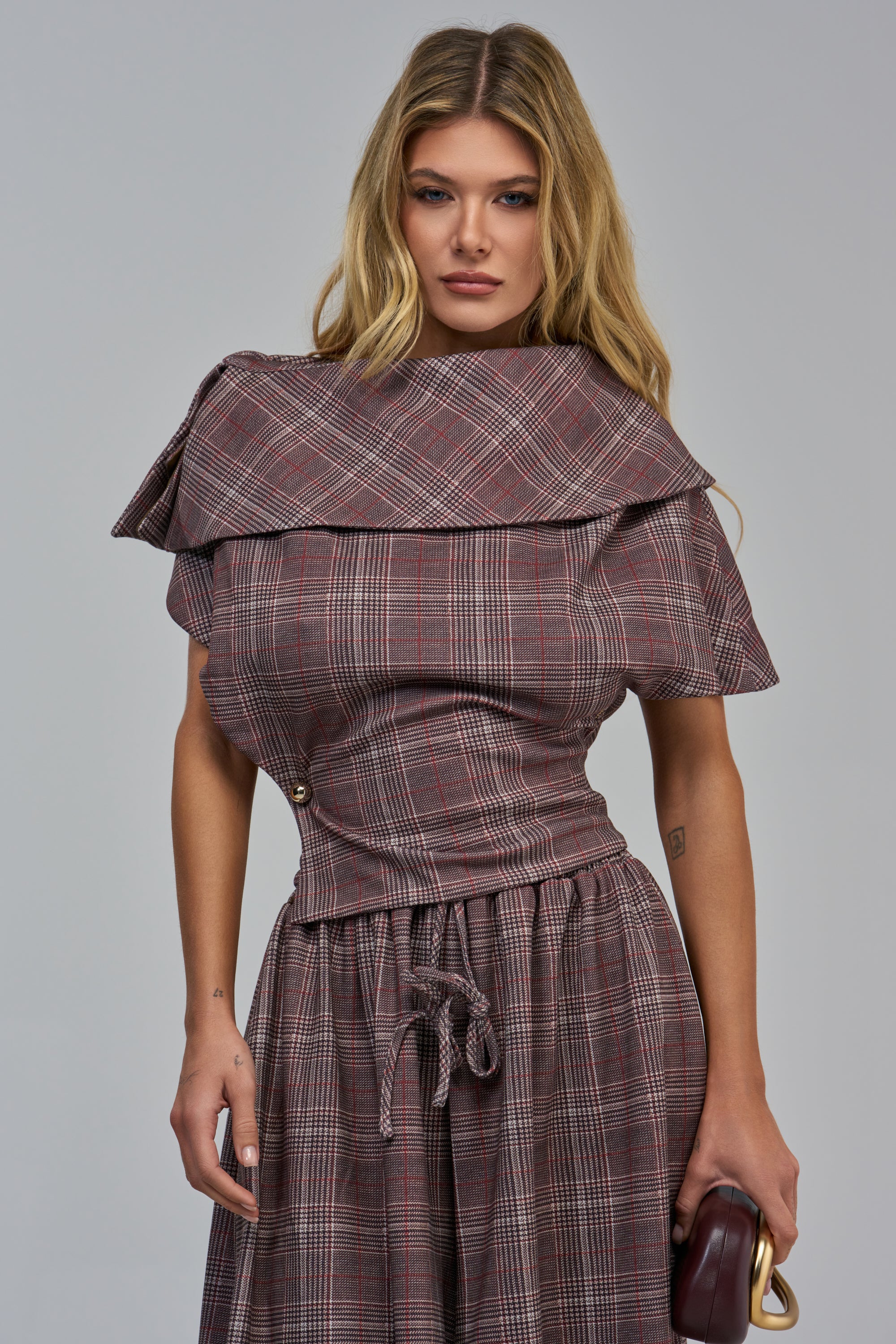A woman in office siren styling wears a brown plaid RUN THE SHOW PLAID TOP with a wide, folded collar and a matching drawstring skirt, holding a small dark brown handbag.