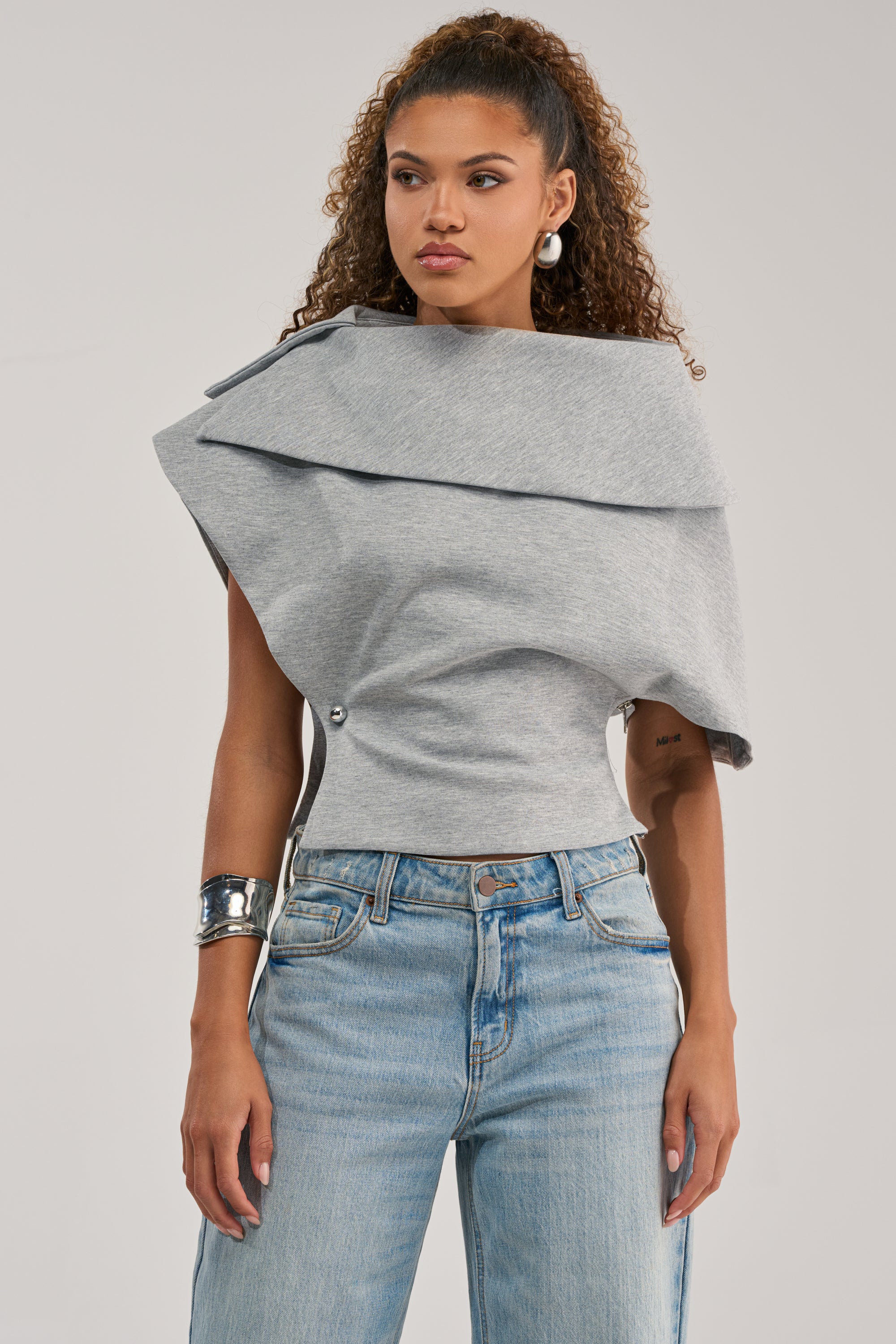 A woman with curly hair, hoop earrings, and a metallic cuff bracelet wears the GIA SCUBA BLOUSE in gray—a sculptural top featuring a wide, folded collar—paired with light blue jeans. She stands with a neutral expression.
