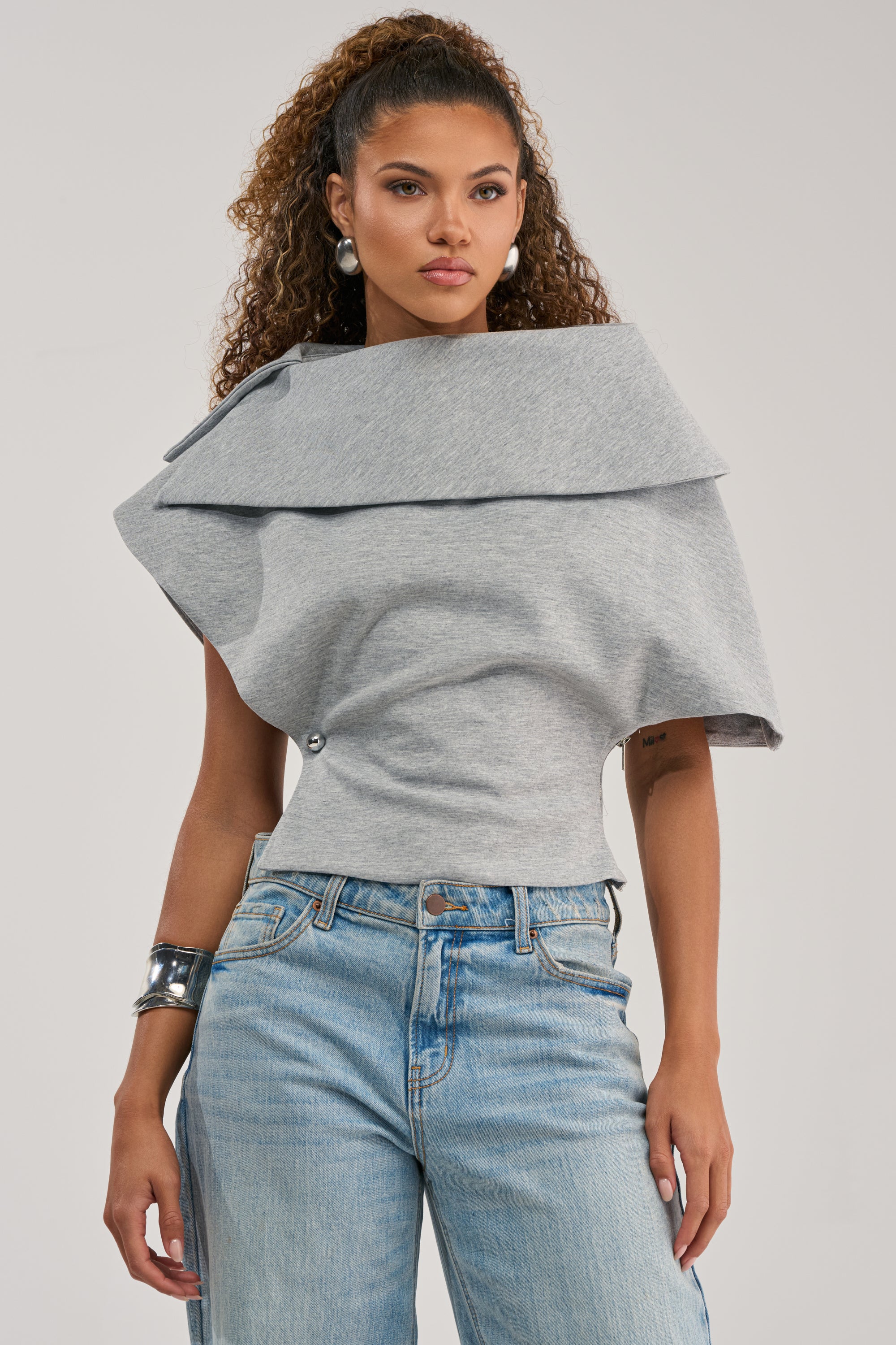 A woman with curly hair wears the GIA SCUBA BLOUSE in gray with light blue jeans, accessorized with large pearl earrings and a silver cuff bracelet, posed against a plain background.