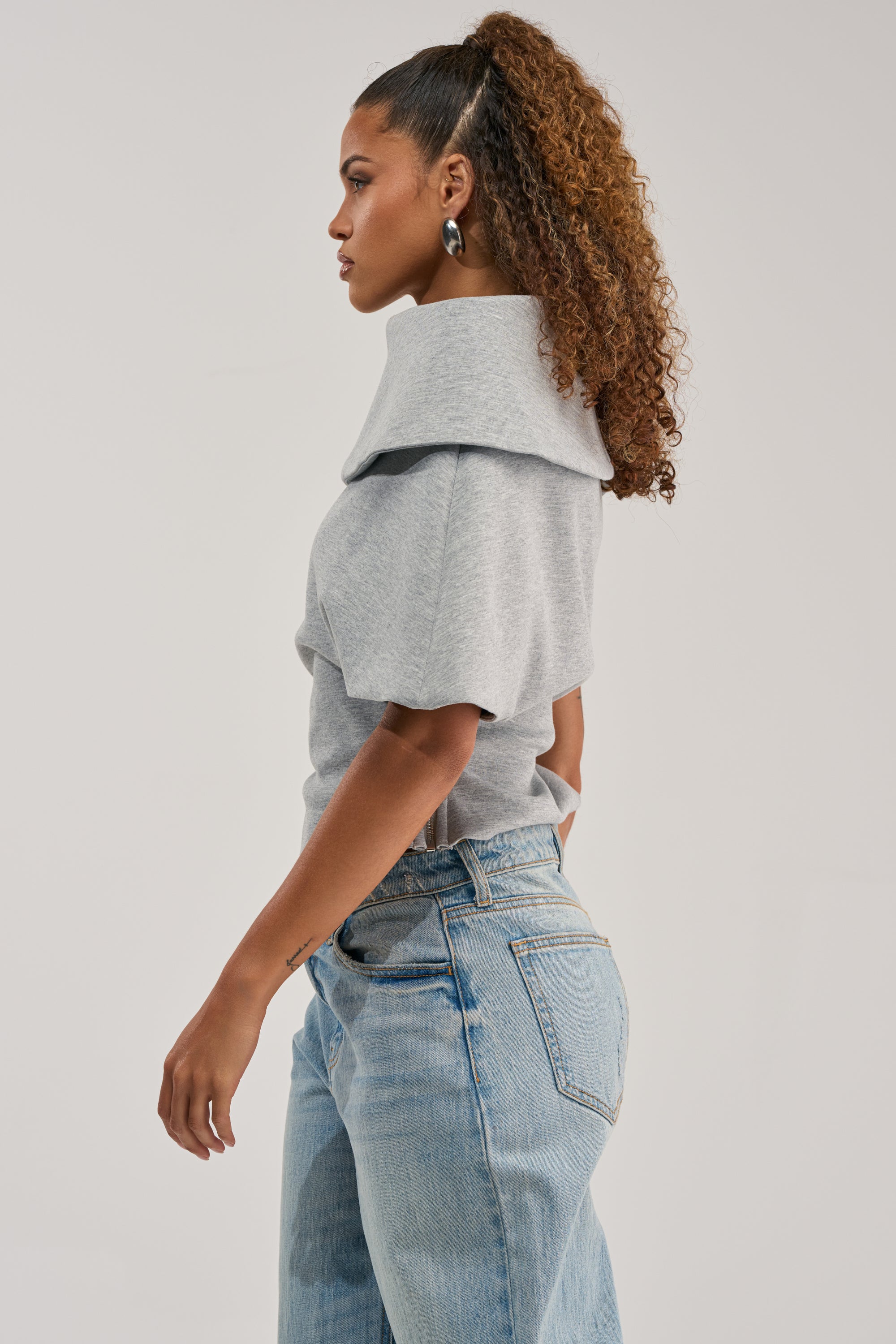 A woman with curly hair styled in a ponytail stands sideways against a plain white background, wearing the GIA SCUBA BLOUSE—a grey top featuring a wide collar—paired with light blue jeans.