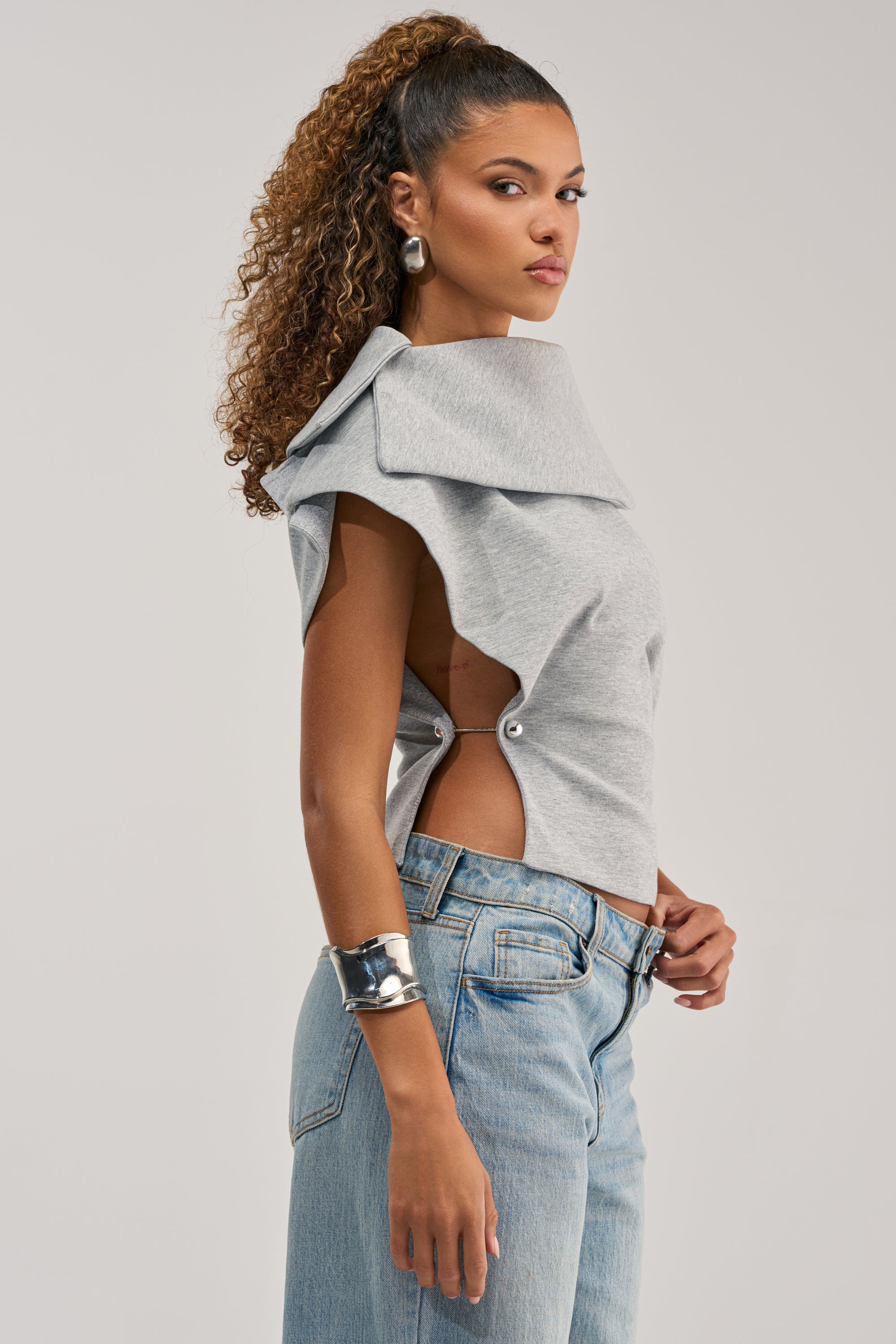 A woman with curly hair in a high ponytail models the GIA SCUBA BLOUSE with side cutouts, blue jeans, large silver hoop earrings, and a wide silver cuff bracelet, posing against a plain background.