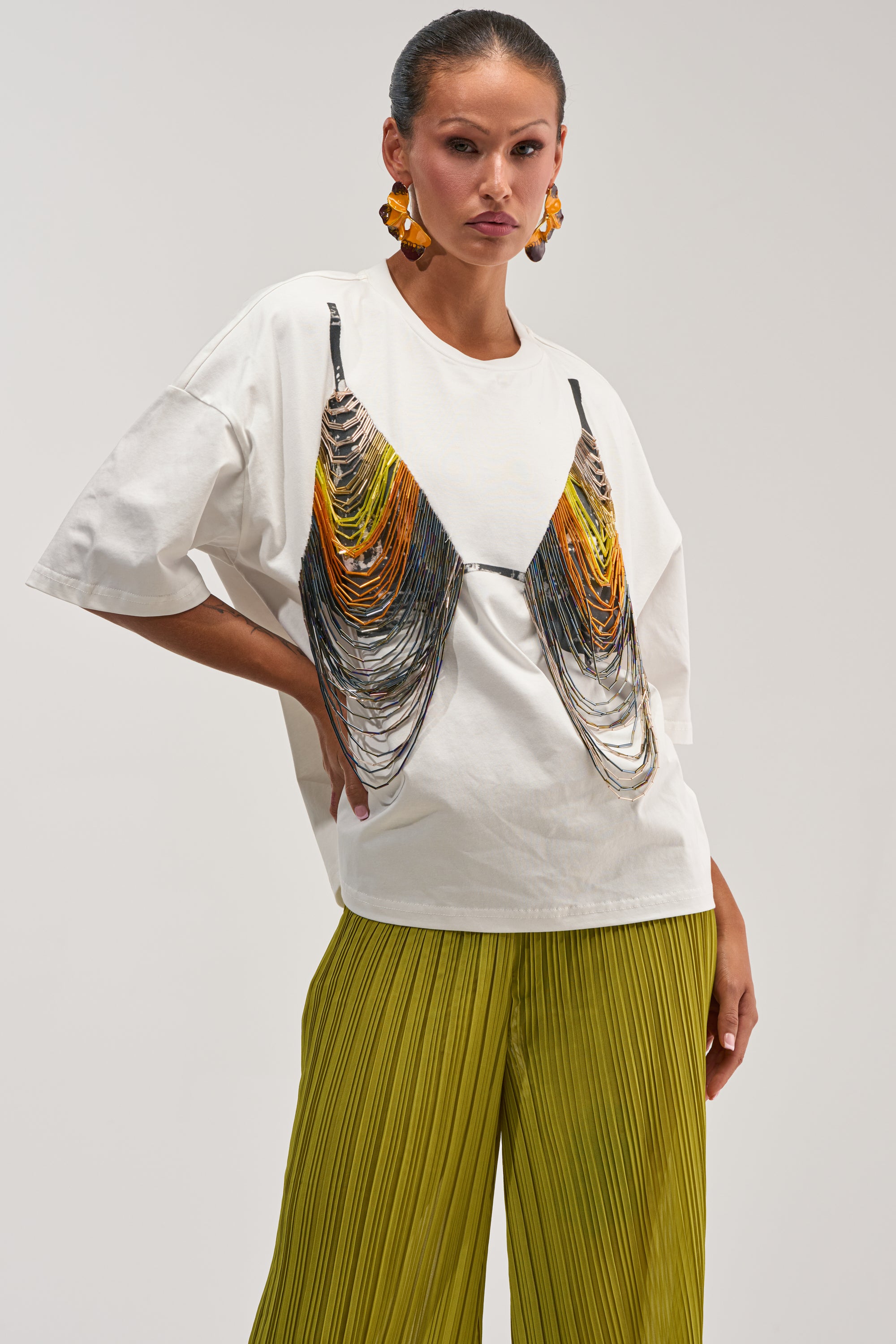Rock the summer streetwear and festival scene in the BEACH VIBES BEADED TEE—an oversized white tee featuring a bold graphic print that mimics a draped, beaded bra in vibrant orange, yellow, and black hues. Styled here with chartreuse pleated pants and statement orange earrings, this eye-catching top is perfect for making a statement wherever you go.