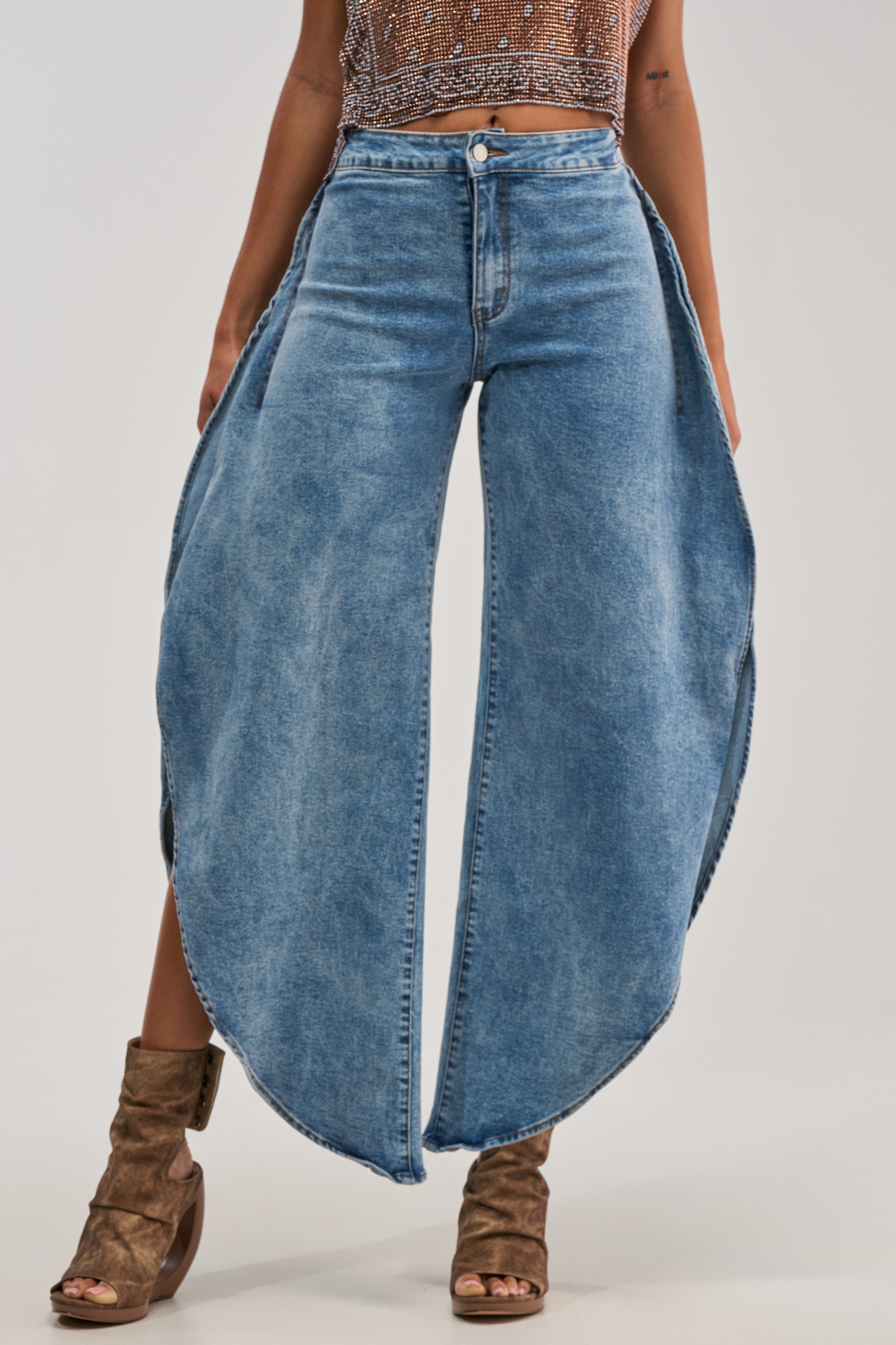 A person is wearing the FEEL THE AIR RELAXED DENIM WITH SIDE OPENING, a pair of high-waisted, light wash jeans featuring extremely wide, curved pant legs, styled with a beaded crop top and brown open-toe heeled boots. The image captures the look from mid-torso to feet.