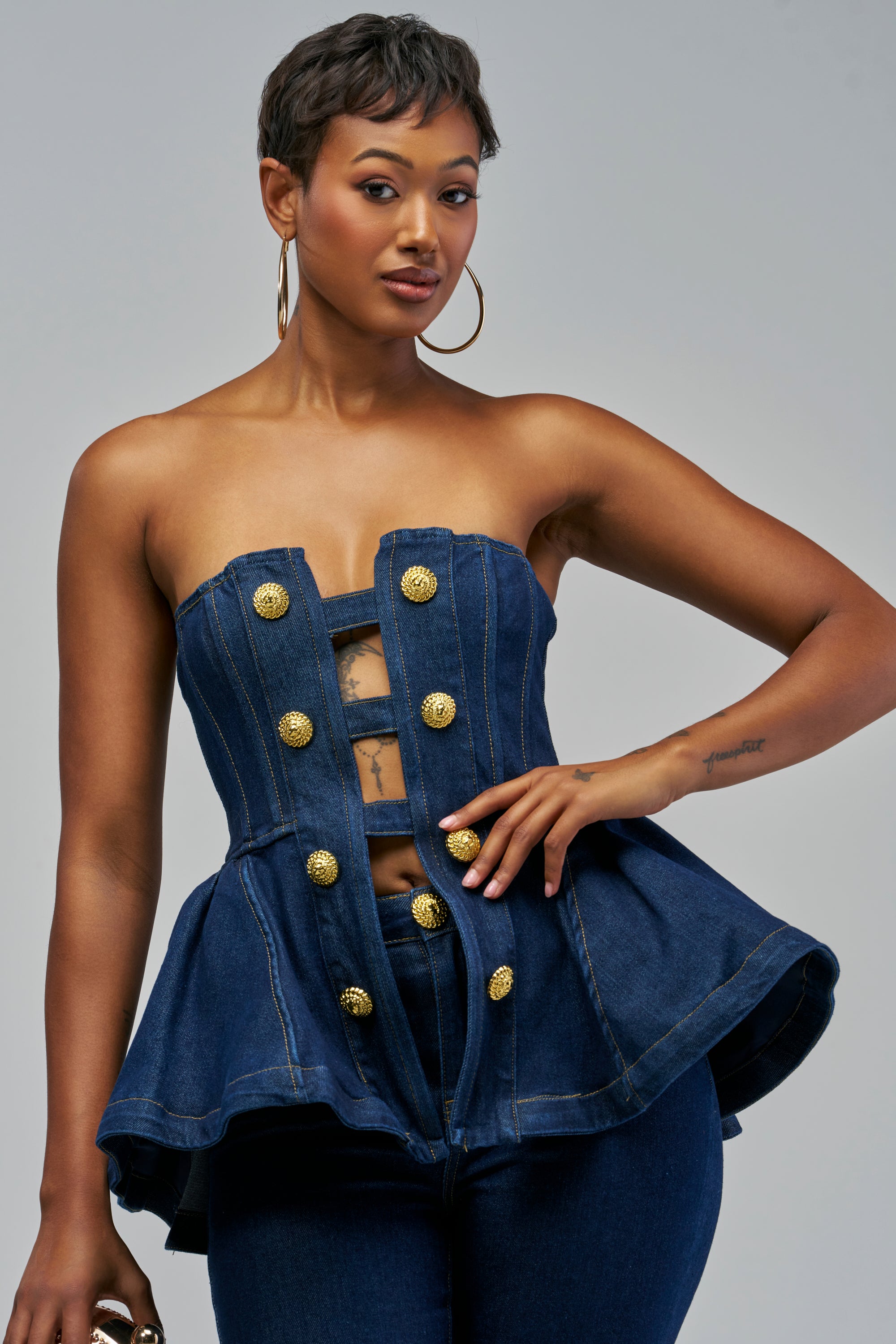 The ANNI DENIM PEPLUM CORSET is a strapless dark denim peplum top featuring a front cutout and gold embossed buttons.