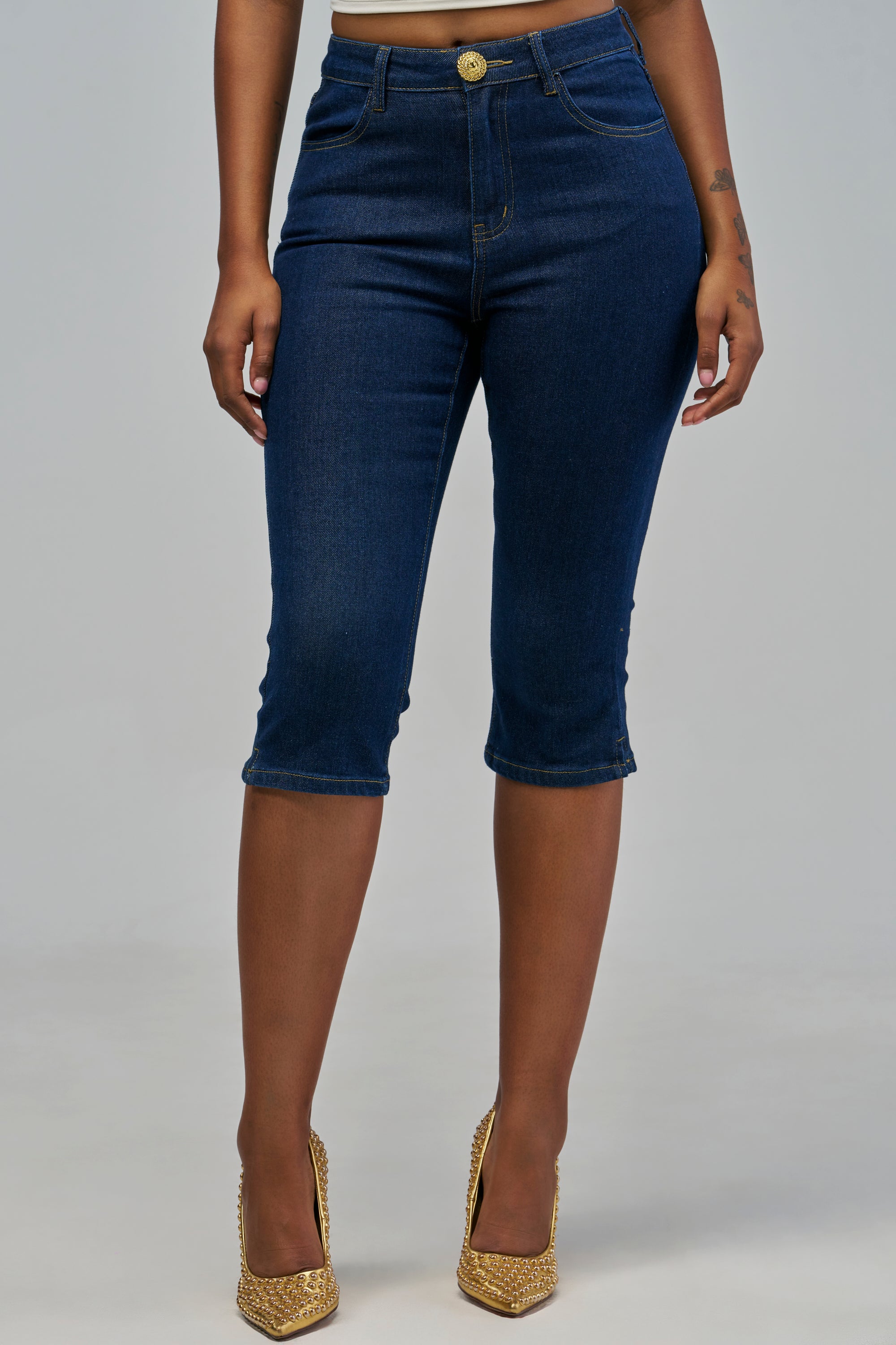 ANNI DENIM CAPRI high-rise dark blue denim capris with a gold button closure, paired with gold studded high heels.