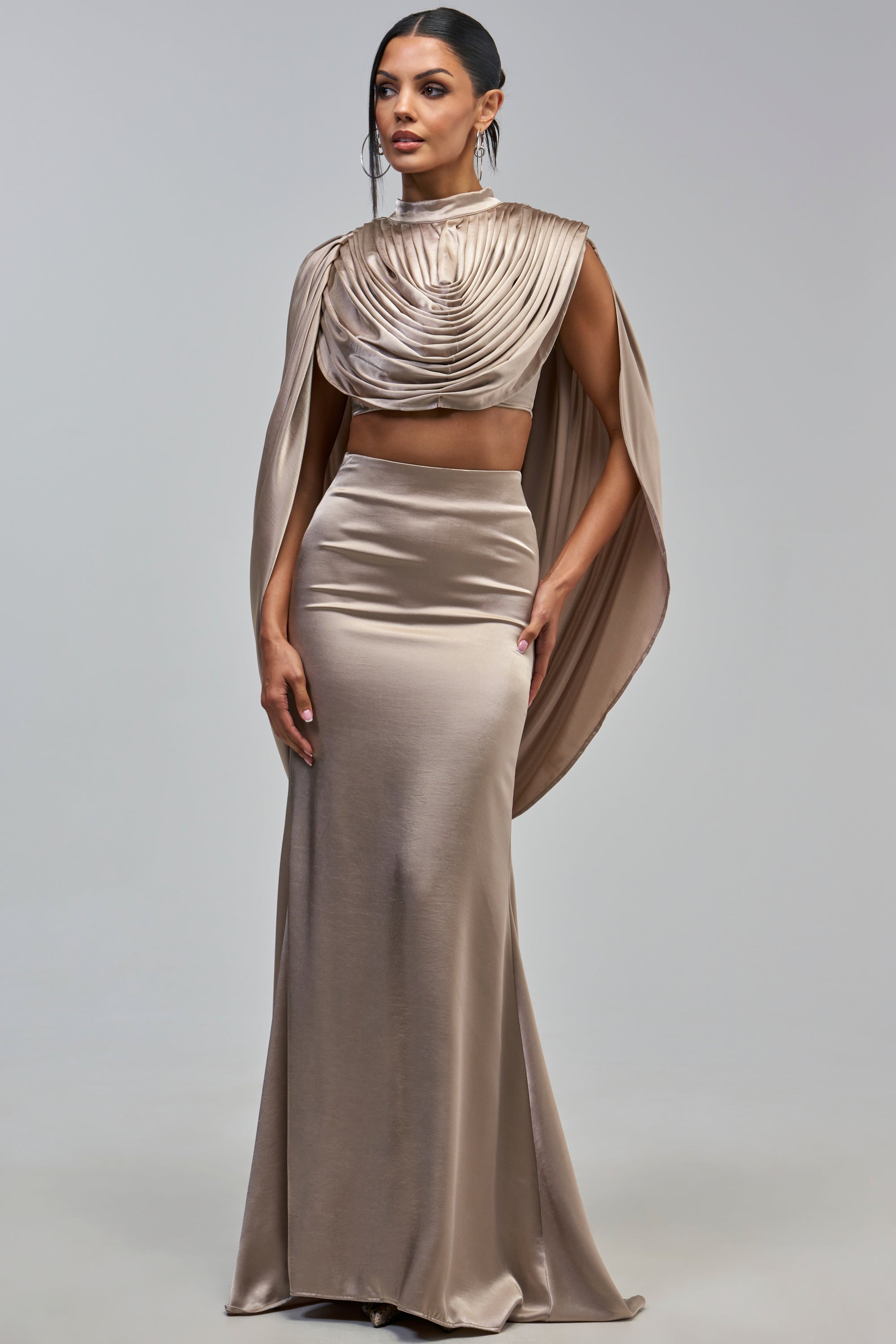 A model wears a champagne-colored high-neck crop top with a draped, cape-style overlay paired with the OPULENCE SATIN MAXI SKIRT in taupe.