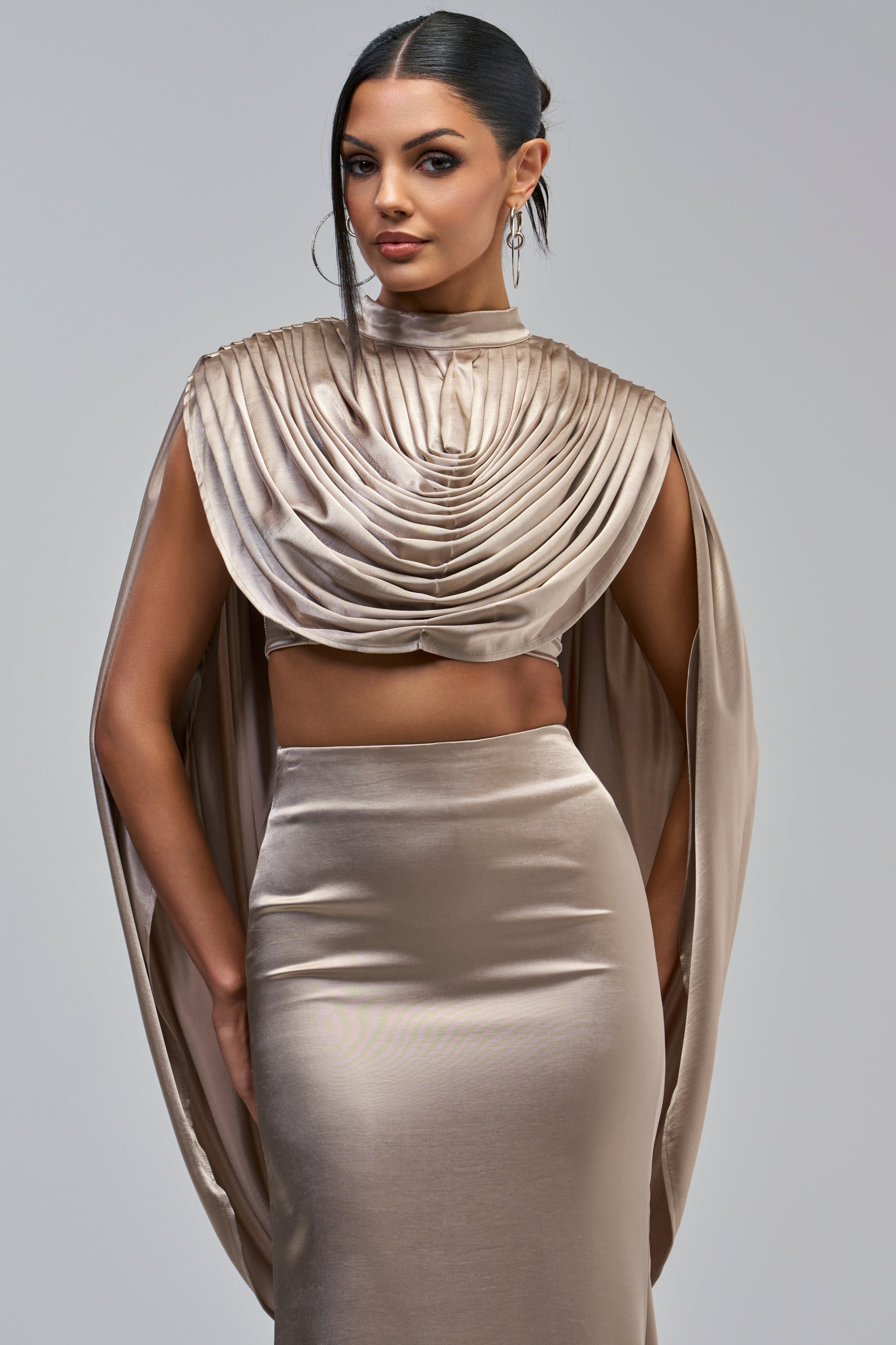 A person wearing an OPULENCE SATIN BLOUSE in taupe, featuring a high-neck, draped cowl neckline, and attached cape sleeves, paired with a matching high-waisted skirt.