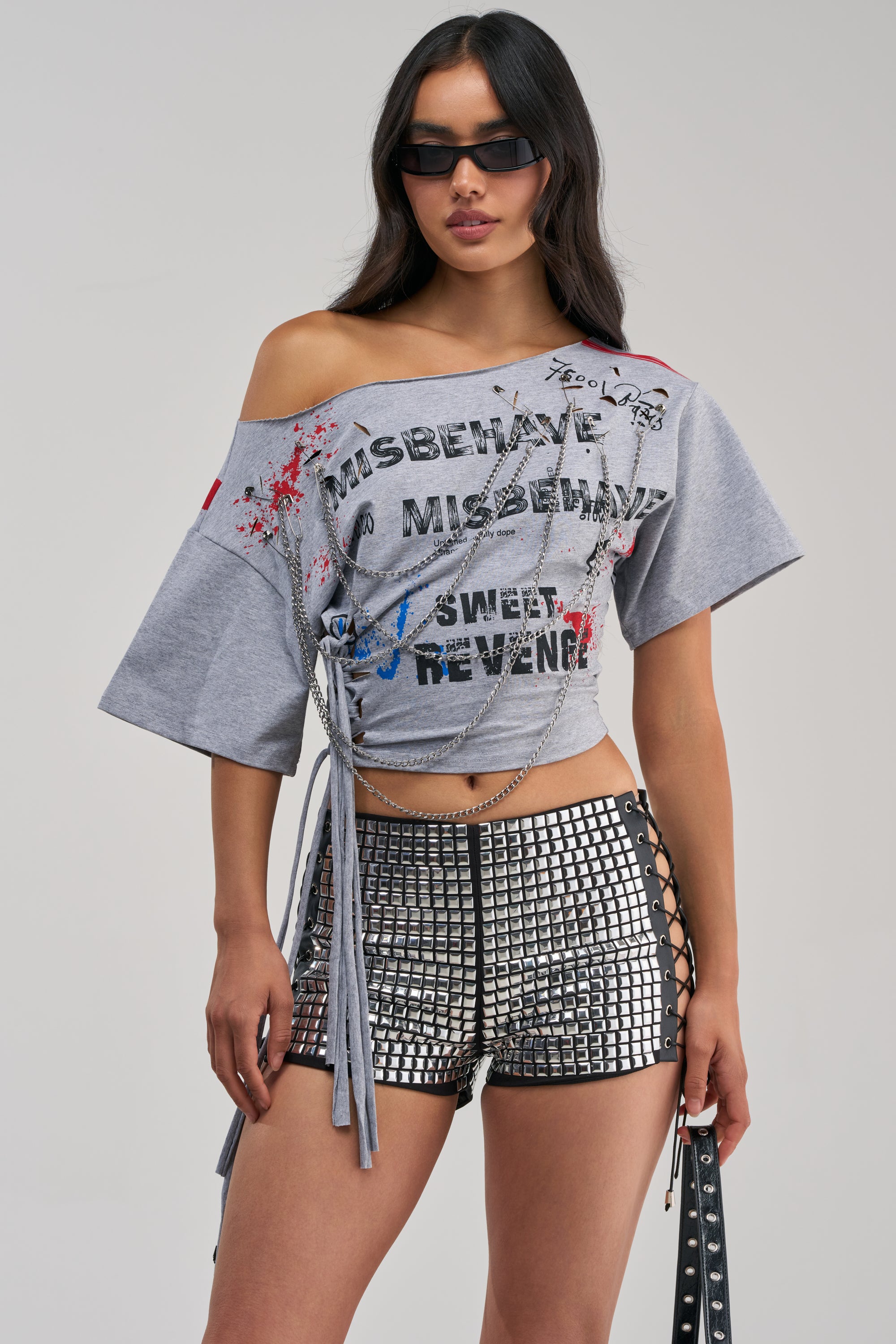 A woman wearing sunglasses and the SWEET REVENGE CHAIN DETAIL TEE—an off-the-shoulder graphic top with text and paint splatters—paired with black-and-white studded lace-up shorts, posing against a plain light background for an effortlessly cool, streetwear-inspired concert look.