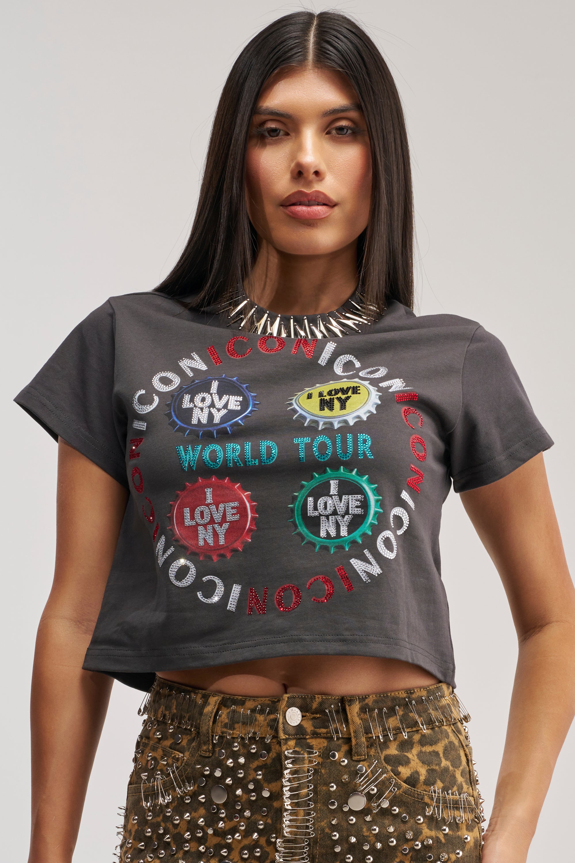 A woman with straight dark hair wears a spiked necklace, the ICONIC GRAPHIC CROPPED TEE that says "Love NY World Tour," and stud-embellished leopard print pants, channeling bold streetwear vibes as she stands against a plain background.