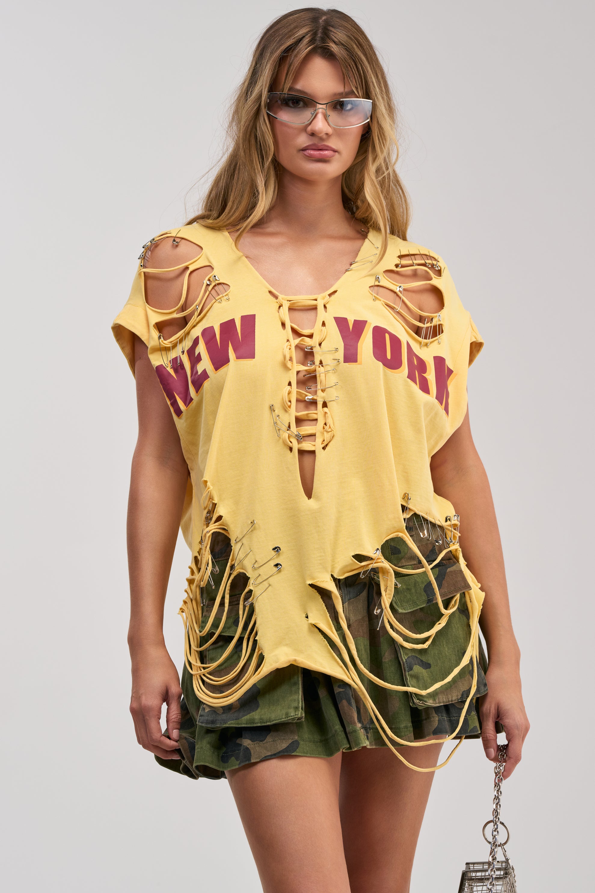 A woman channels streetwear vibes in the VARSITY STAR DISTRESSED TEE, a yellow graphic top adorned with a bold “New York” design, laces, and safety pins. She completes her festival-ready look with green camo shorts, sunglasses, and a small silver chain bag.
