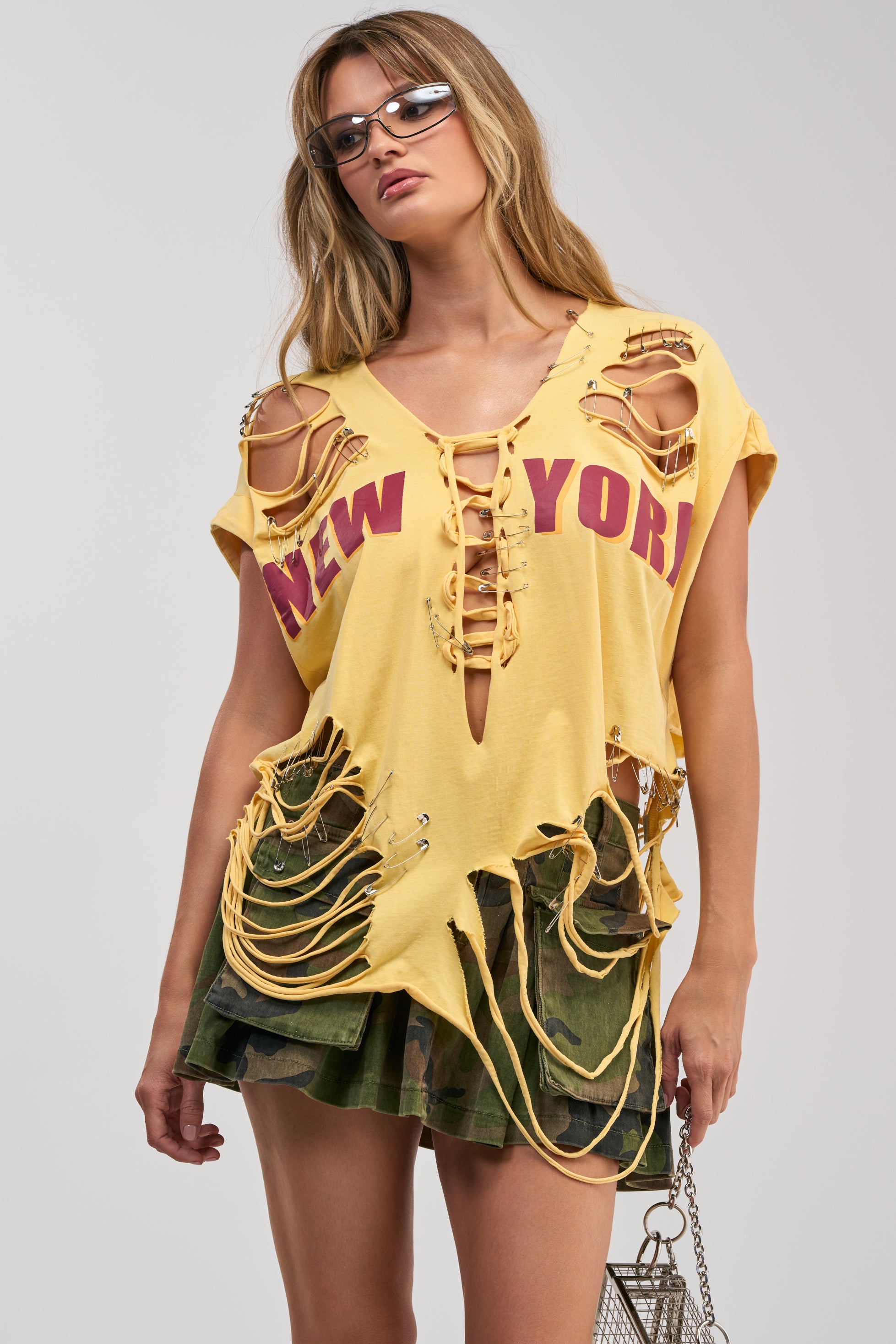 Wearing sunglasses, a yellow VARSITY STAR DISTRESSED TEE featuring “New York” lettering and a lace-up front, paired with a short camouflage skirt and carrying a small silver bag—a perfect look for streetwear festival styling.