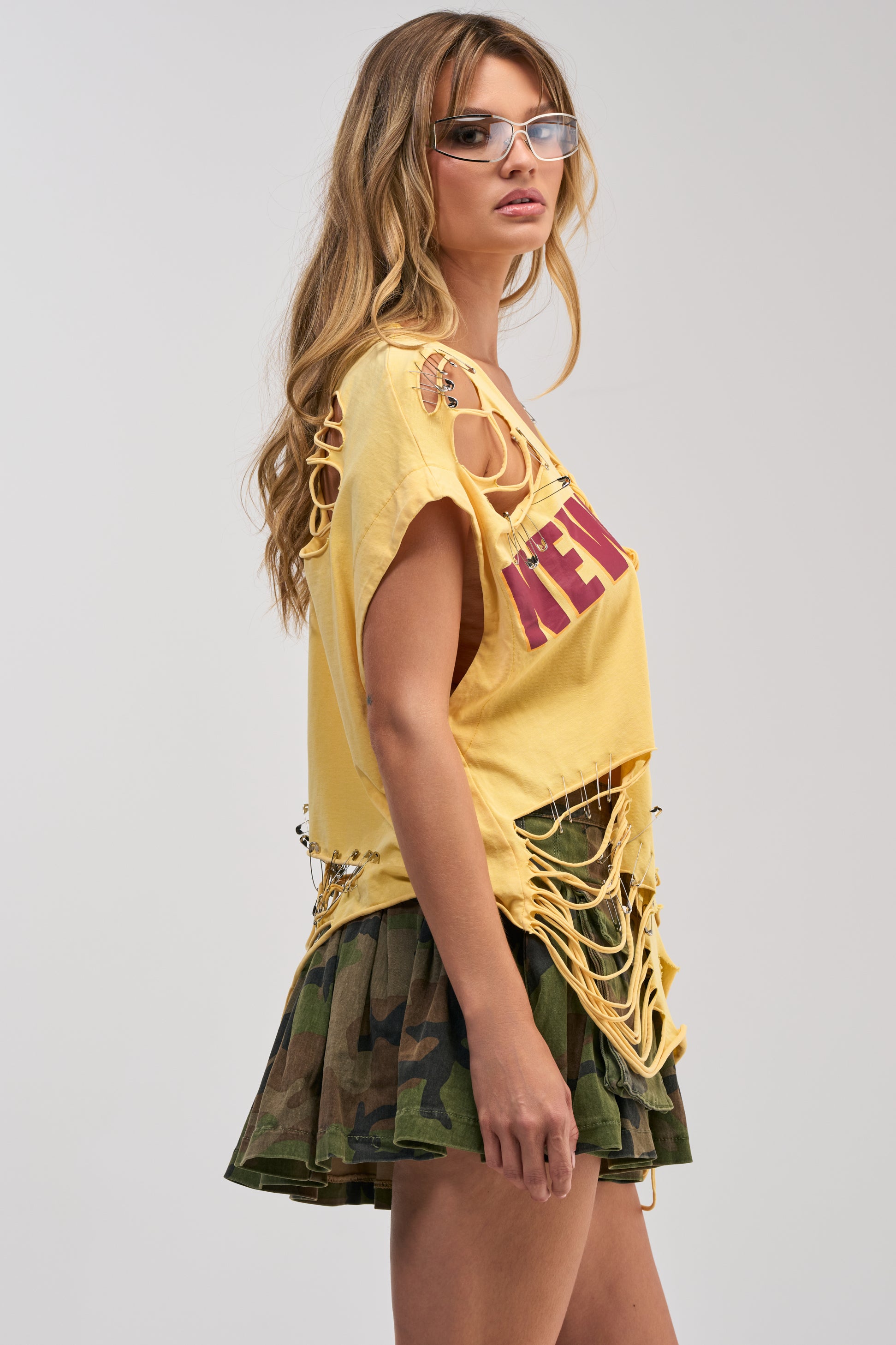 A woman wearing clear glasses, the VARSITY STAR DISTRESSED TEE—a yellow graphic top with cutouts and chain accents—and a camouflage pleated skirt stands against a plain background, showcasing bold festival styling inspired by streetwear influences.