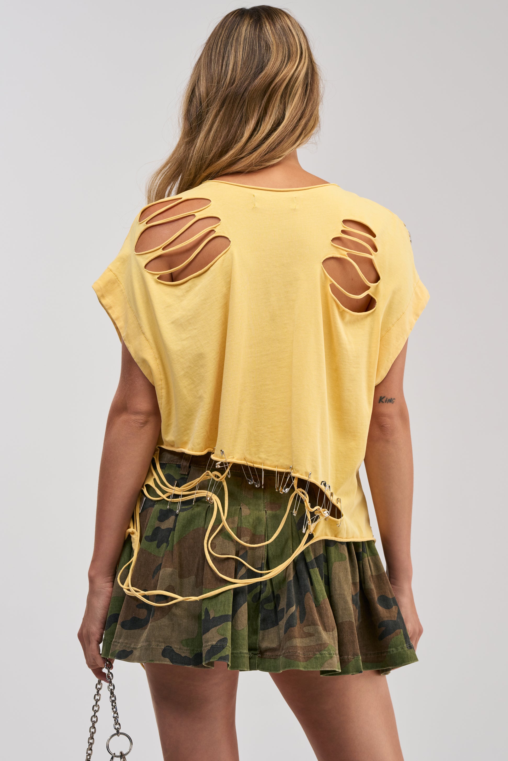 A person with long wavy hair wears the VARSITY STAR DISTRESSED TEE, a yellow graphic top featuring large back slits and hanging strings, paired with a pleated camouflage mini skirt and holding a chain strap. The effortless festival-inspired look stands out against a plain light gray background.