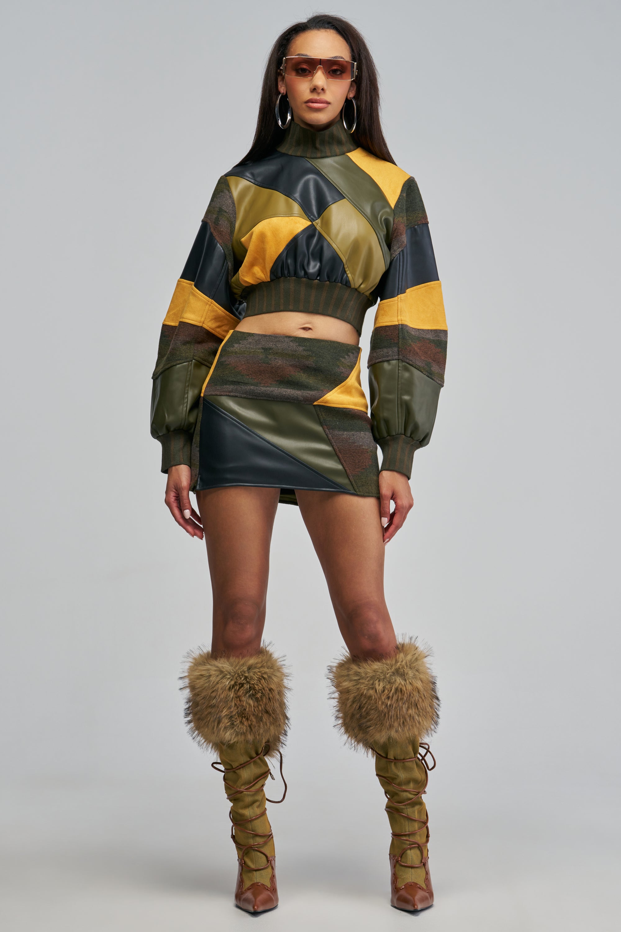 A woman wears a fall patchwork crop top and the IMOGINE PATCHWORK SKIRT in shades of olive, black, and yellow, paired with lace-up boots featuring faux fur cuffs.
