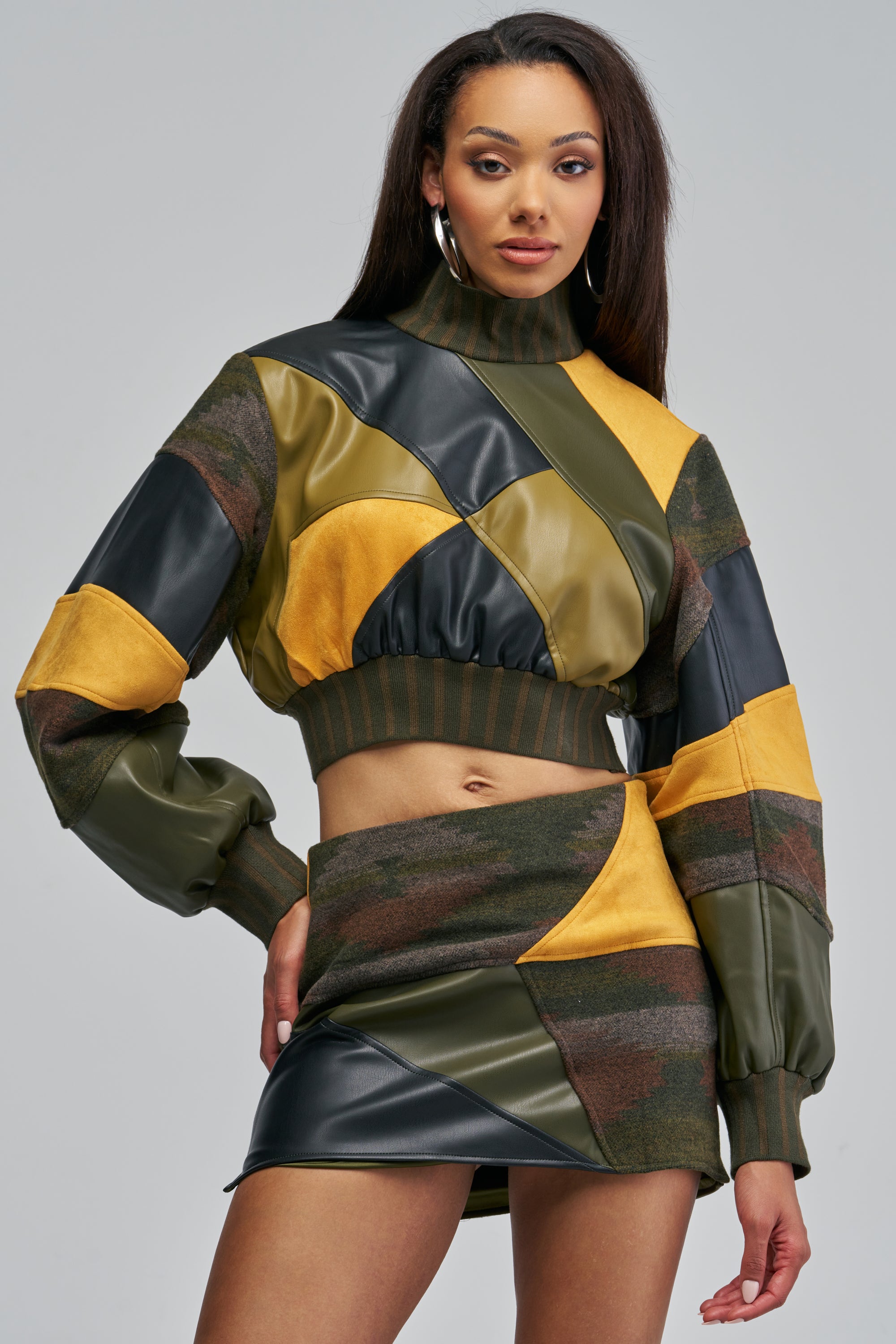 A woman wears an IMOGINE PATCHWORK SWEATSHIRT and matching mini skirt, both featuring a patchwork design of olive green, mustard yellow, black, and camouflage fabric in a versatile fall styling.