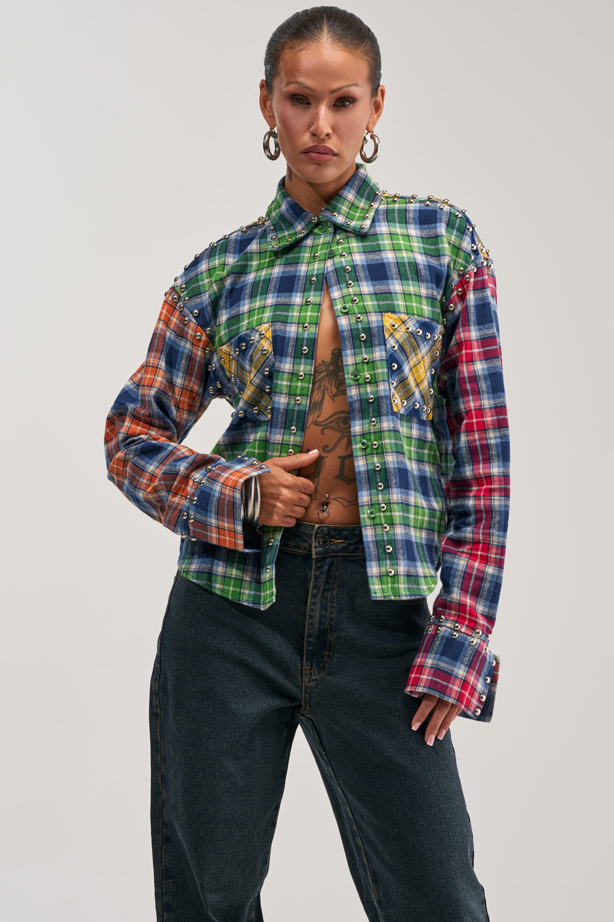 Wearing the JUST FOR FUN PLAID EMBELLISHED BUTTON DOWN—featuring vibrant patchwork plaid and studded details, styled partially unbuttoned to reveal a chest tattoo—and paired with dark jeans, the individual stands confidently against a plain backdrop, showcasing bold streetstyle flair.