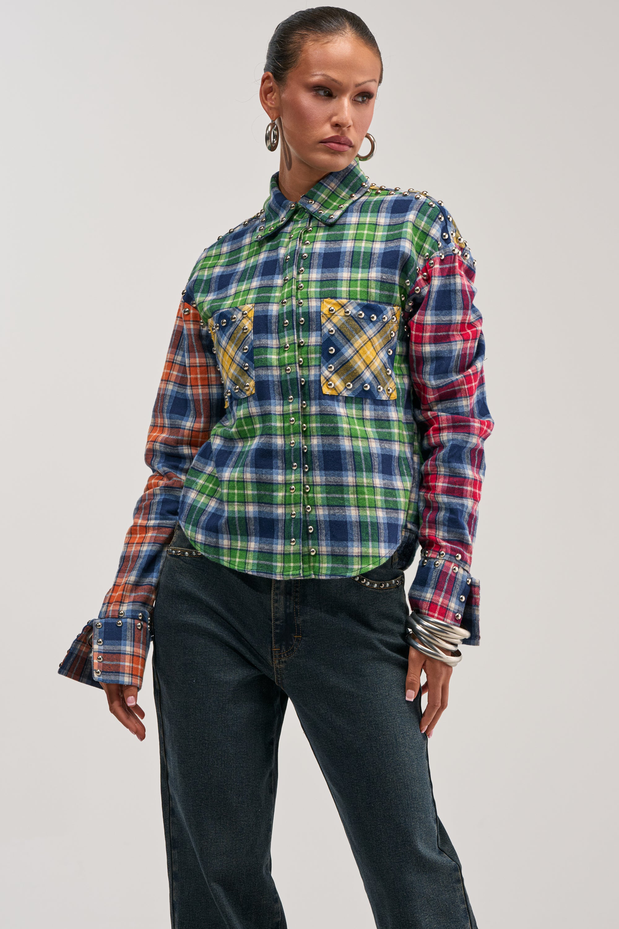 Wearing the JUST FOR FUN PLAID EMBELLISHED BUTTON DOWN with vibrant plaid pattern and stud accents, paired with dark jeans, this person poses confidently with one hand on their hip against a plain background for a cool streetstyle look.