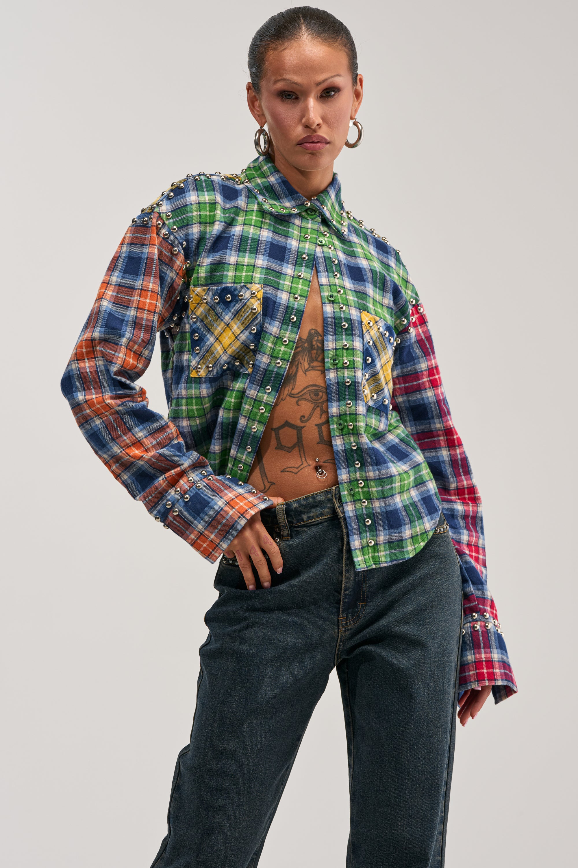 Wearing the JUST FOR FUN PLAID EMBELLISHED BUTTON DOWN—a multicolored patchwork plaid shirt adorned with pearl accents—paired with dark jeans and showcasing a tattooed torso, this person stands confidently against a plain backdrop for a striking streetstyle look.