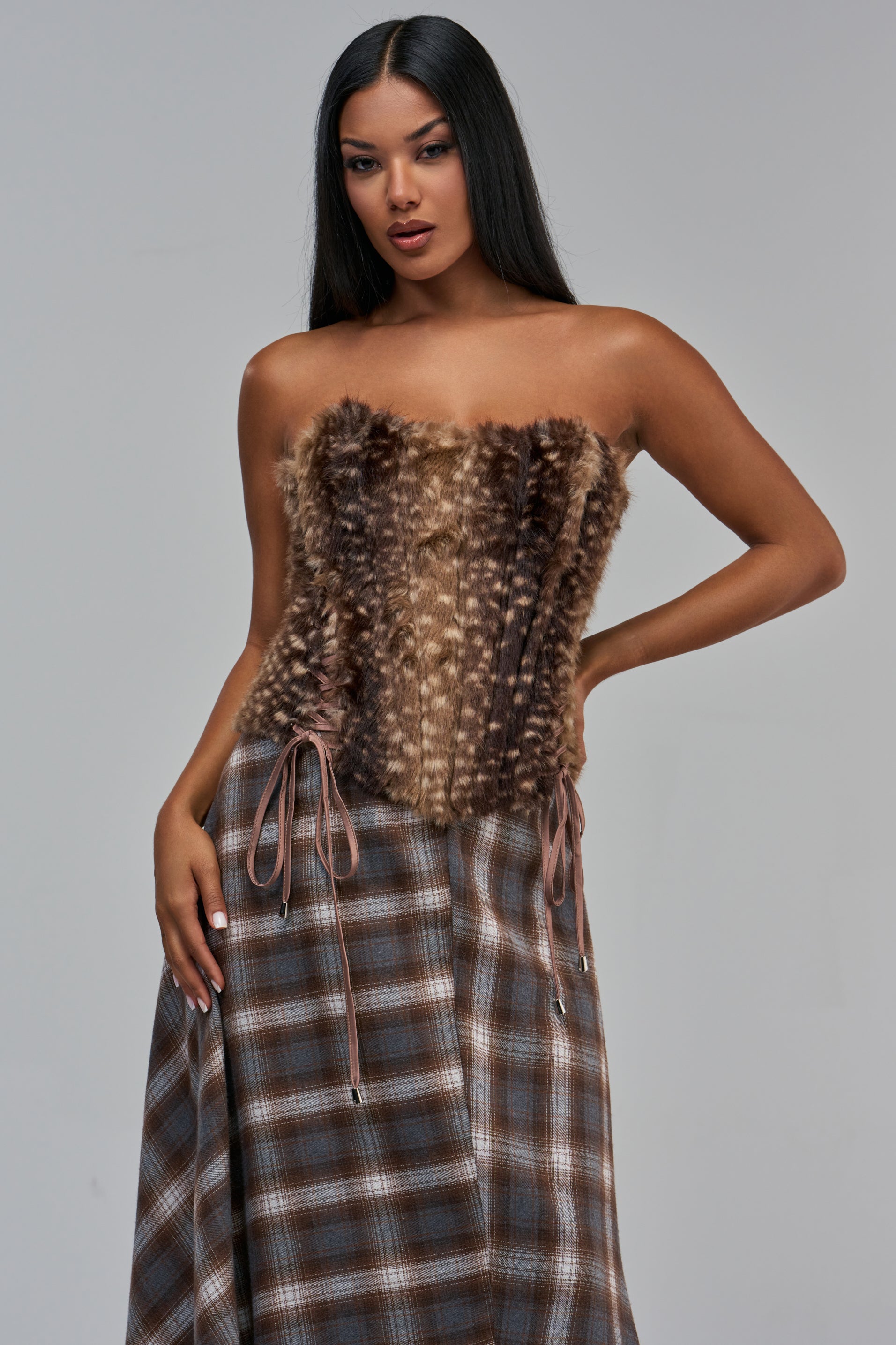 A model wearing a brown BAMBI BABE FAUX FUR CORSET with side lace-up details and a brown plaid skirt.
