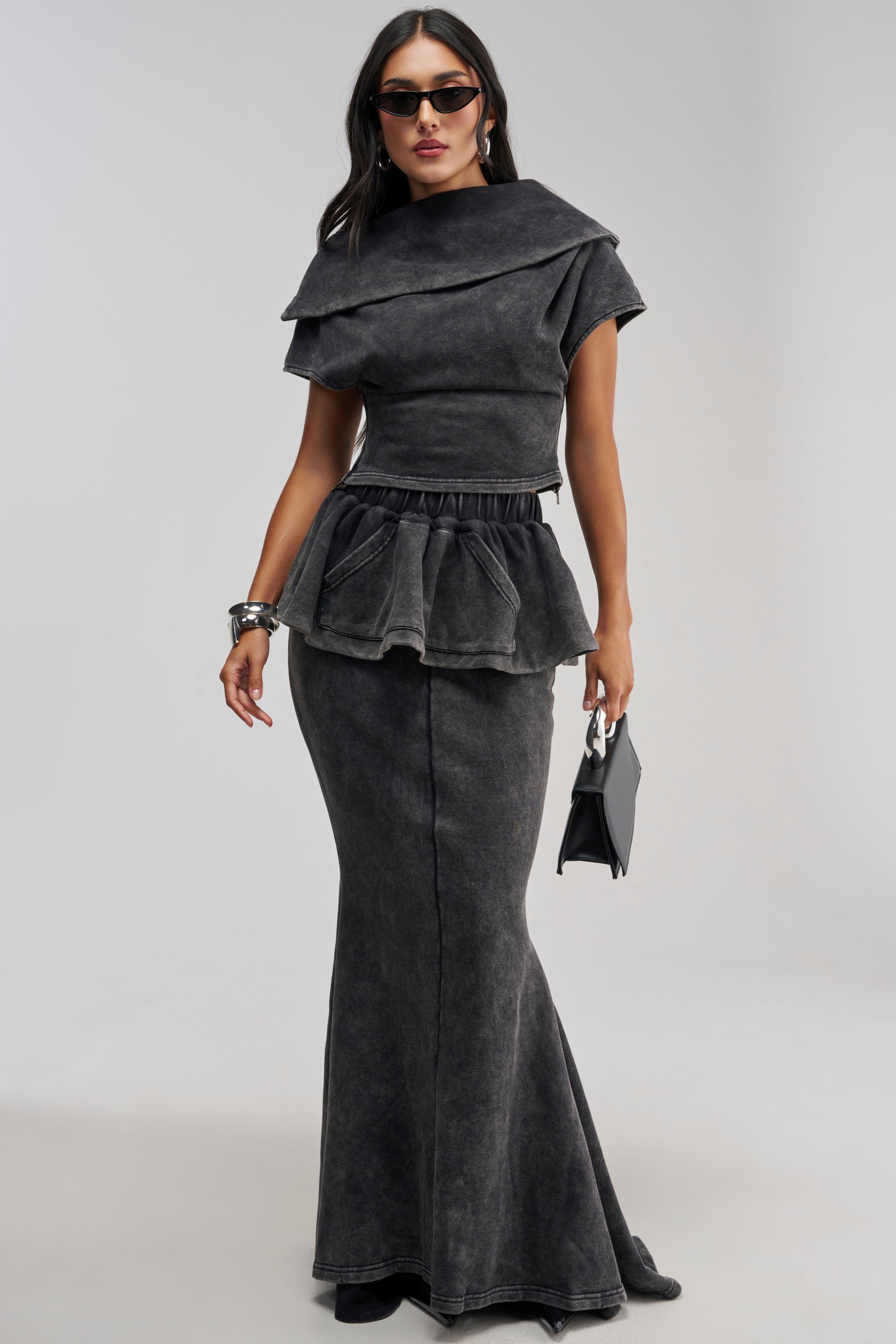 Wearing the JOCELYN MINERAL WASHED MAXI SKIRT set, a woman styles her look with sunglasses, a small black handbag, and bracelets, radiating effortlessly chic streetwear vibes against a plain light background.