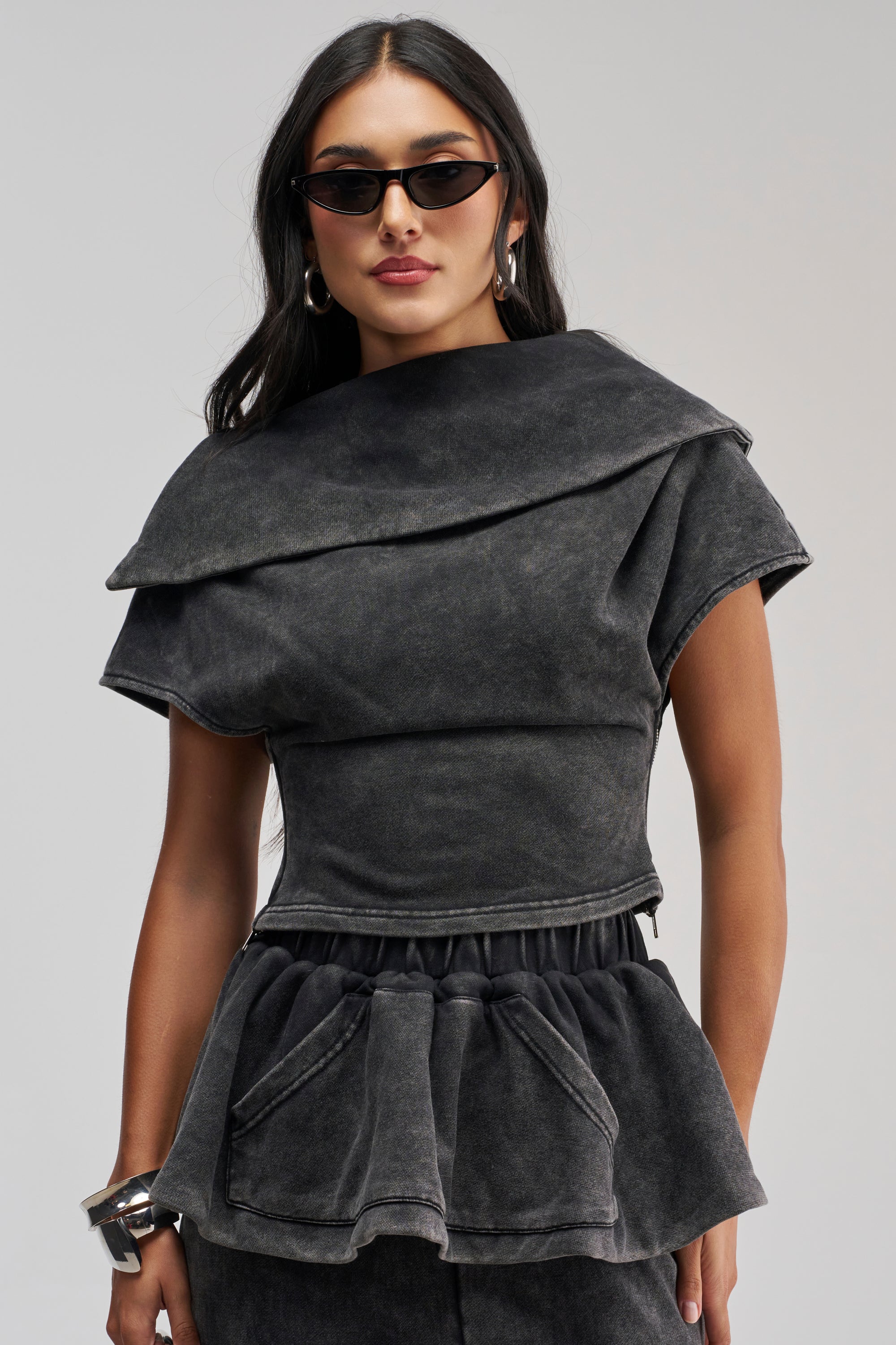 Woman wearing black angular sunglasses and the JOCELYN MINERAL WASHED SWEATSHIRT, featuring a structured, layered dark gray sculpted fit with asymmetrical short sleeves and peplum detail, standing against a plain background.