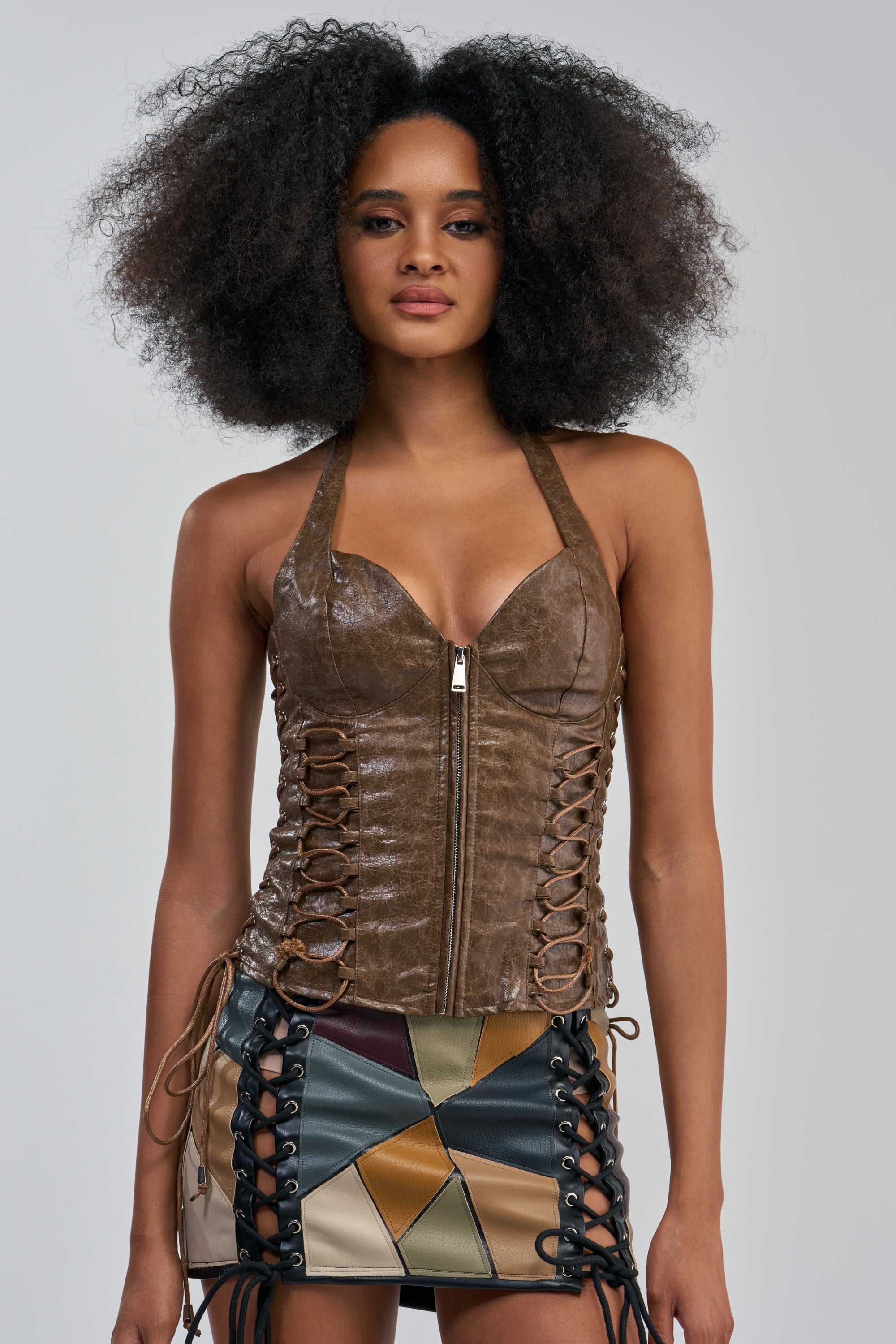 A model with voluminous curly hair showcases the FIGHT FOR ME PU CORSET TOP IN BROWN, which features textured lace-up details in a brown faux leather finish. Styled as a bold concert look, it is paired with a matching multi-colored patchwork mini skirt with side lacing, set against a plain grey background.