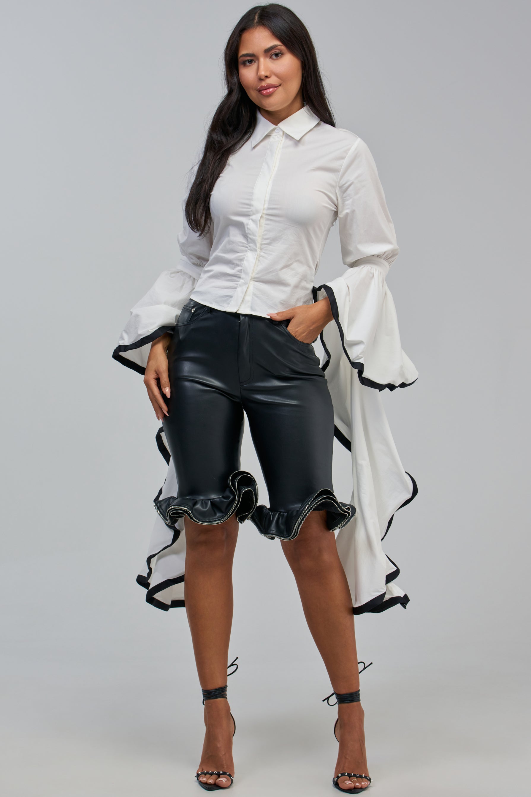 Wearing the ETHEREAL FLARE SLEEVE BLOUSE styled as a standout workwear piece, this woman showcases its dramatic ruffled sleeves and timeless collared button-down design. She pairs the blouse with black knee-length shorts featuring ruffled hems and completes the look with black high-heeled sandals, standing against a plain background.