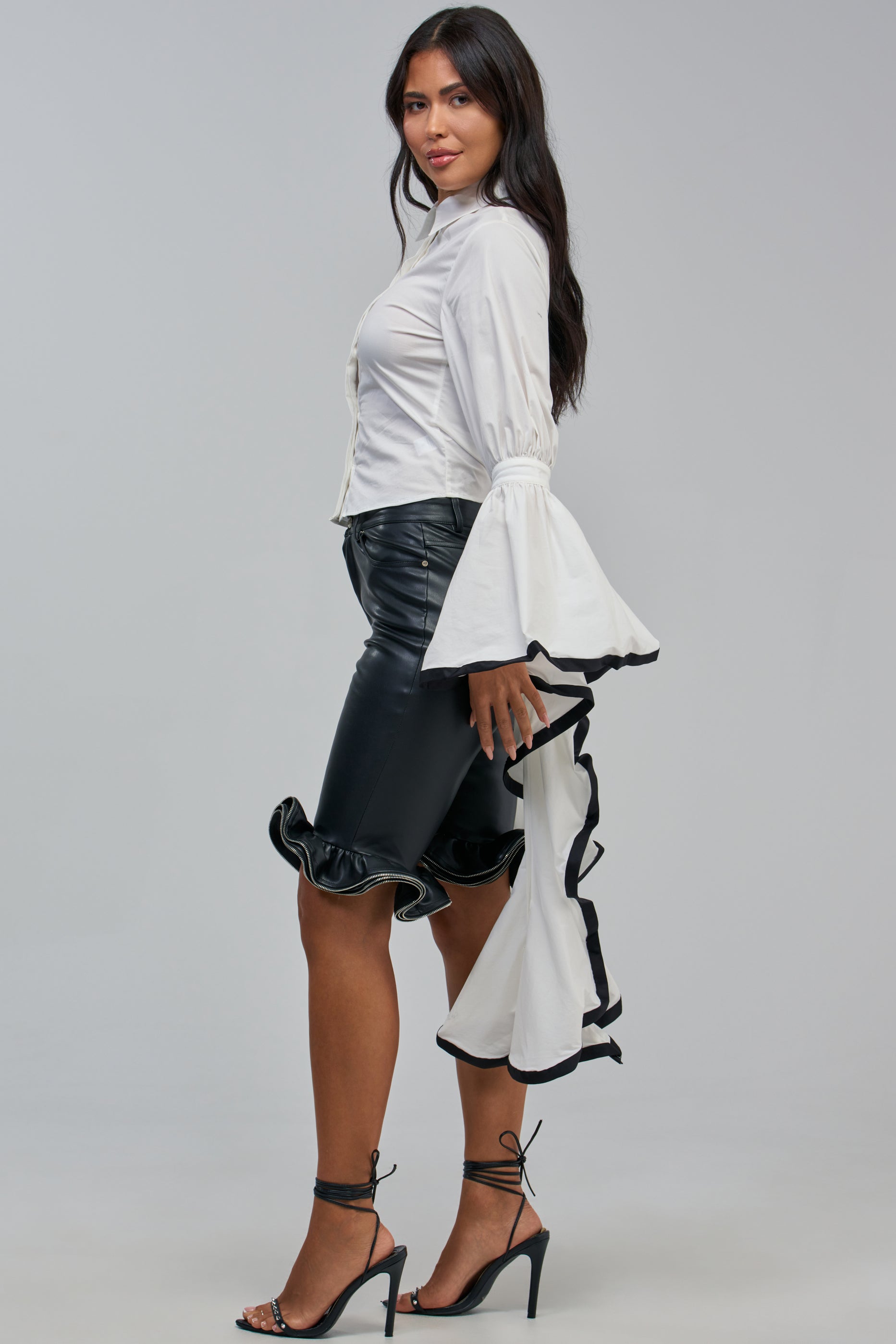 Woman in the ETHEREAL FLARE SLEEVE BLOUSE—a striking workwear top with dramatic flared cuffs, a collared button-down front, and an elongated back detail—paired with a black leather skirt featuring a ruffled hem and black strappy heels, standing in profile against a plain gray background.
