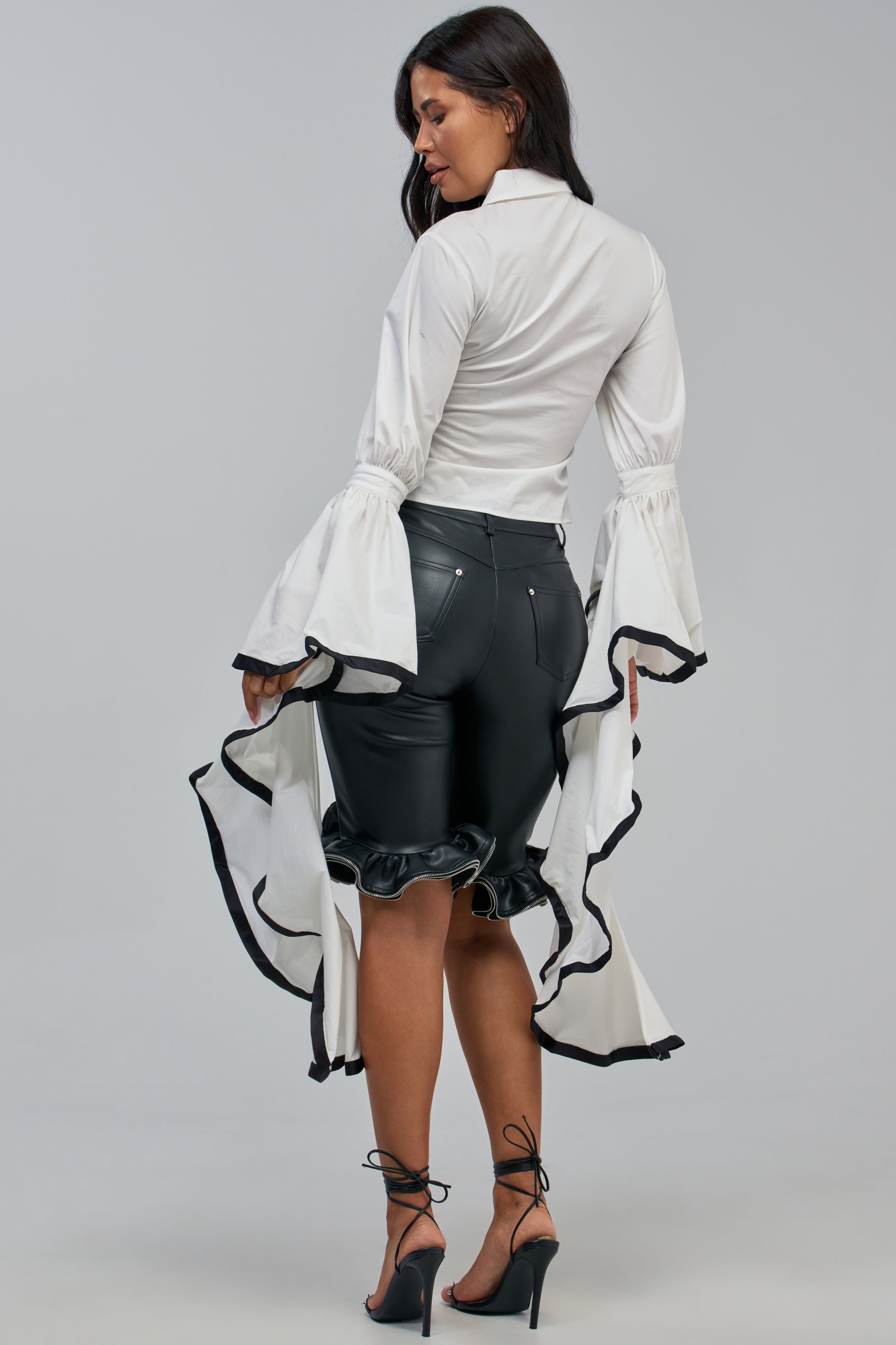 A woman wears the ETHEREAL FLARE SLEEVE BLOUSE—distinguished by its dramatic flared ruffle sleeves and long ruffled back—styled as a statement workwear top with black high-waisted leather pants and black high-heeled sandals, set against a plain gray background.