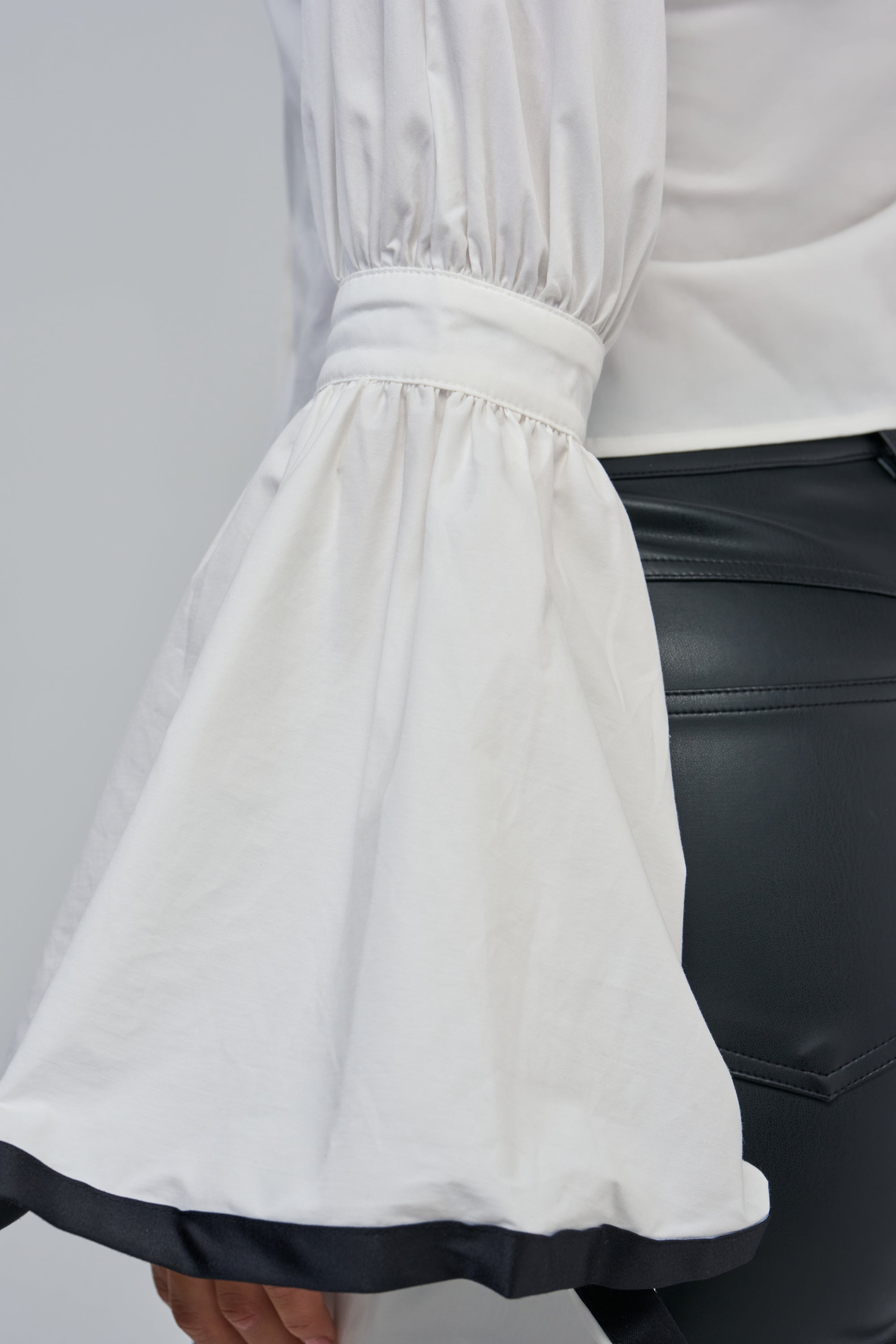 A close-up of a person wearing the ETHEREAL FLARE SLEEVE BLOUSE, a white top with wide, flared ruffle sleeves and gathered detailing near the wrist, paired with black leather pants. This statement workwear piece adds an elegant touch to the modern look.