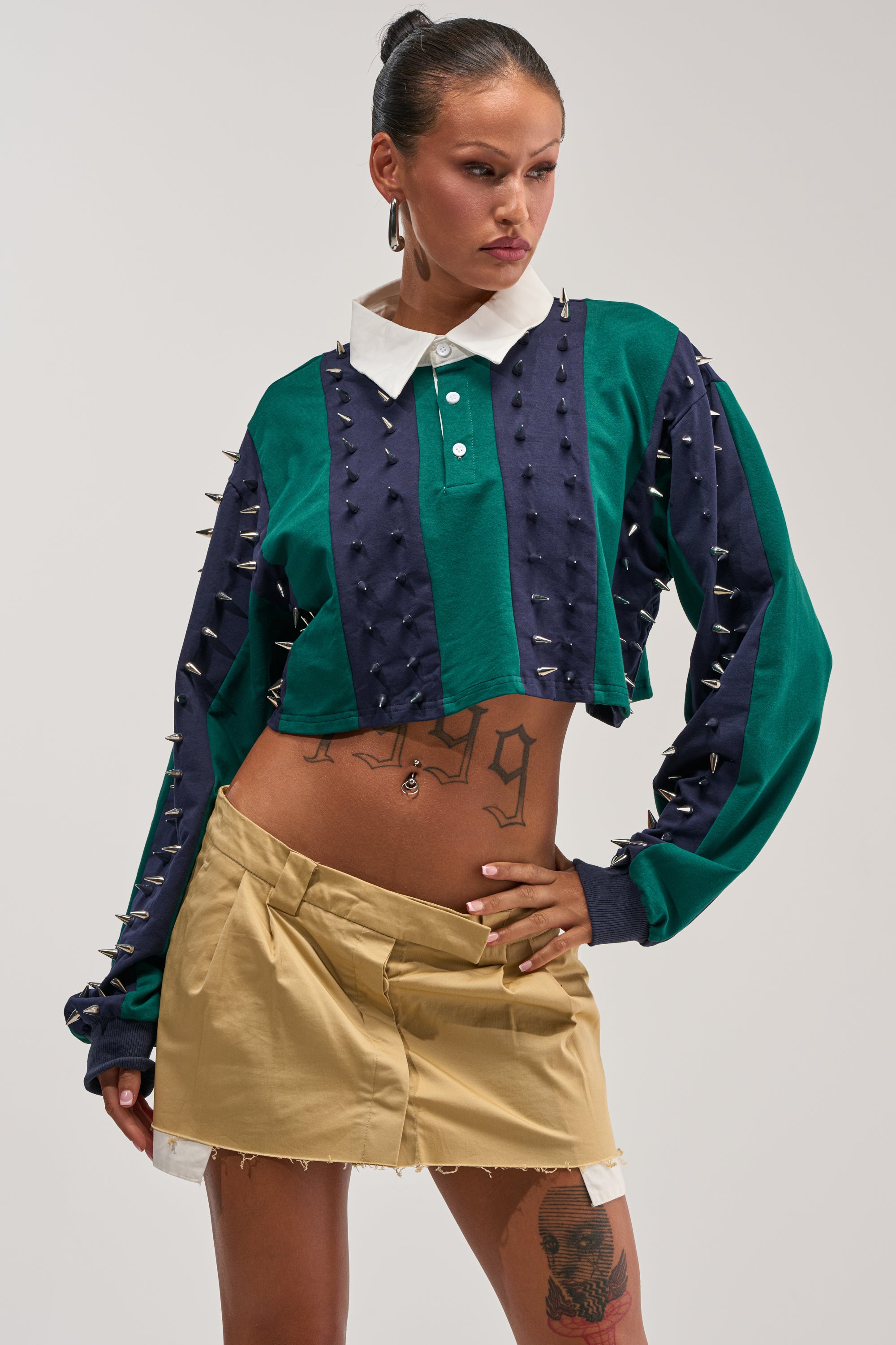 A woman wears the DONT TOUCH CROPPED POLO SHIRT—a green and navy rugby-style top with silver spike accents—paired with a beige mini skirt and showcasing her tattoos as she poses confidently against a plain background, embodying bold streetwear vibes.