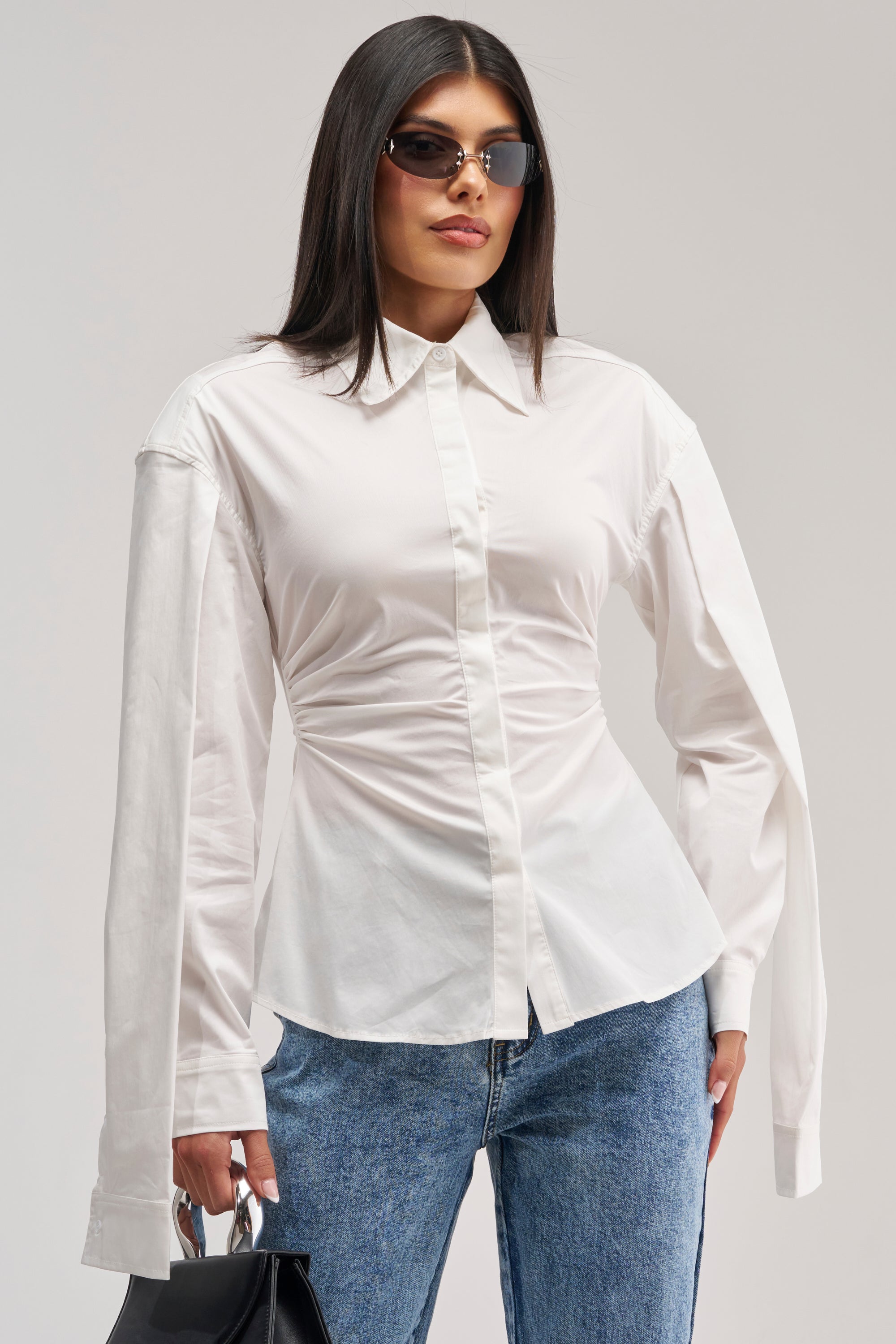 A woman poses against a plain background, wearing sunglasses, blue jeans, and the BOSS UP POPLIN BLOUSE—a fitted white poplin blouse with extra-long sleeves—and holding a black handbag.