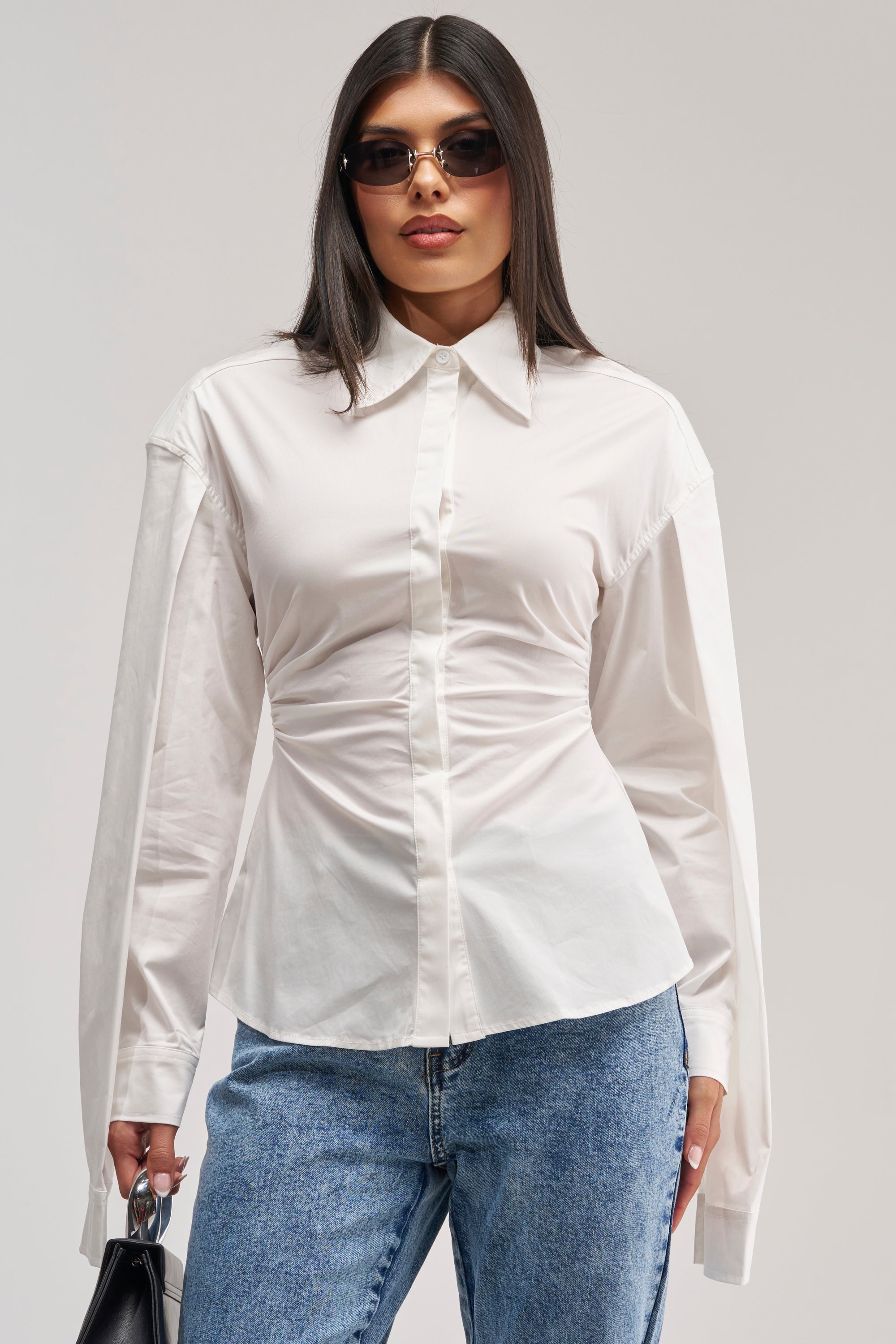 A woman with straight dark hair wearing sunglasses, blue jeans, and holding a small black handbag stands against a plain background, styled in the BOSS UP POPLIN BLOUSE with long sleeves.