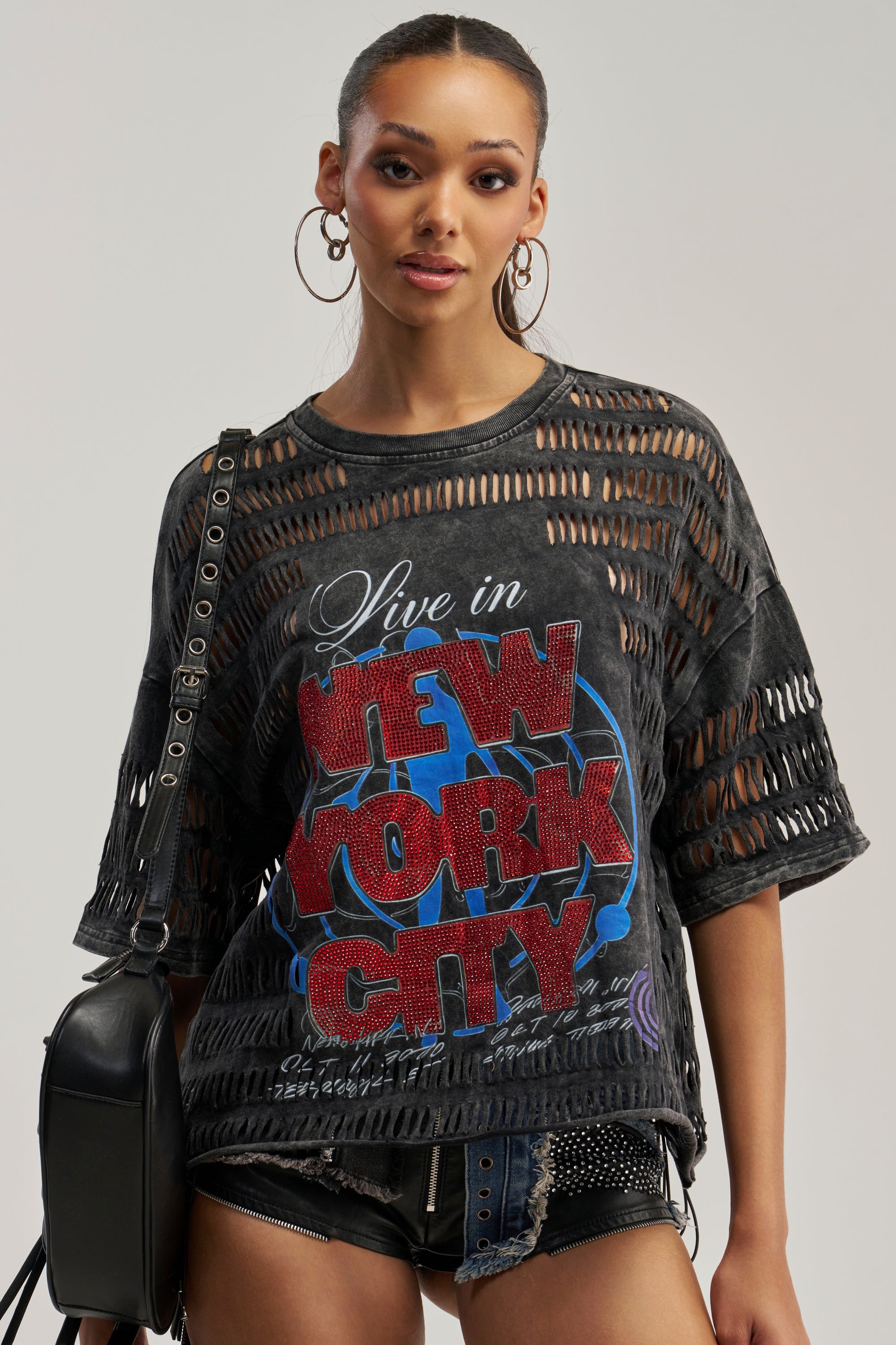 A woman wearing large hoop earrings, a grungy graphic NYC RHINESTONE DISTRESSED TEE with a paneled, distressed design, and studded shorts poses with a black shoulder bag against a plain background.