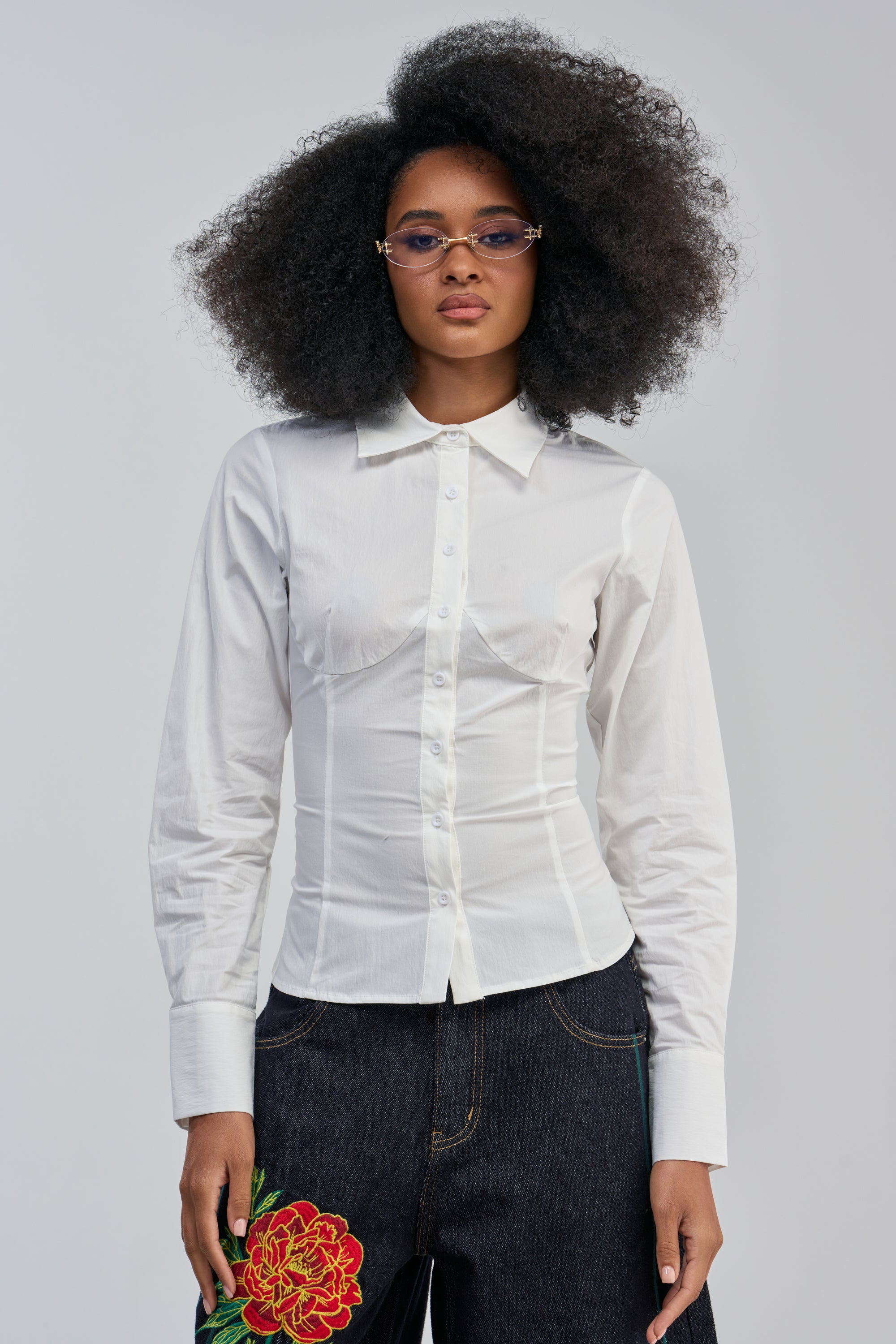 Here’s a rewrite of your sentence using the provided product data: A woman with natural curly hair wears the OFFICE SIREN POPLIN BLOUSE in white, styled with dark jeans featuring a red floral design. She accessorizes with stylish glasses and stands against a plain background.
