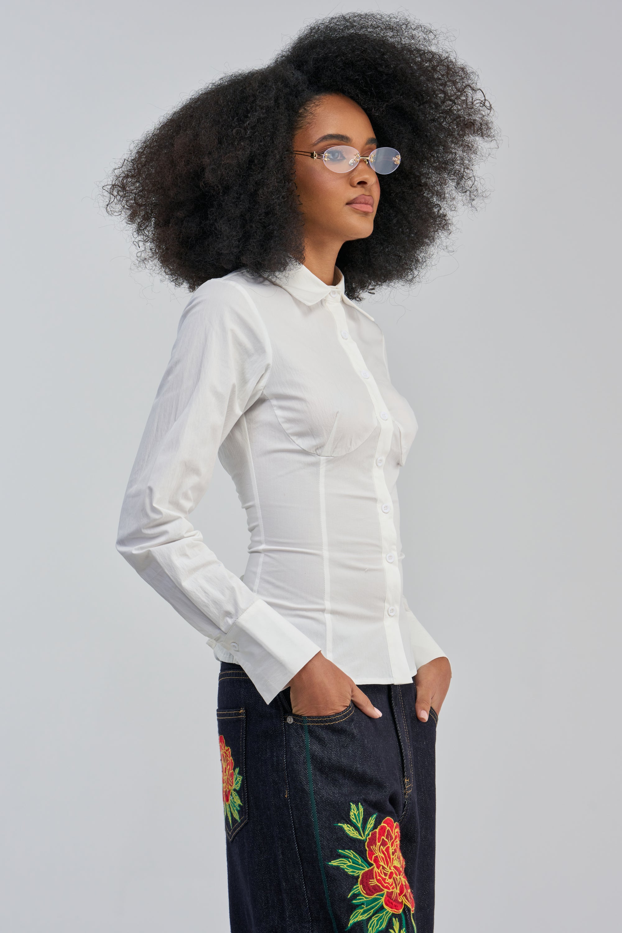A woman with natural curly hair and small glasses stands in profile against a plain light background, wearing the OFFICE SIREN POPLIN BLOUSE in white—sculpted fit—and dark jeans featuring colorful floral embroidery.