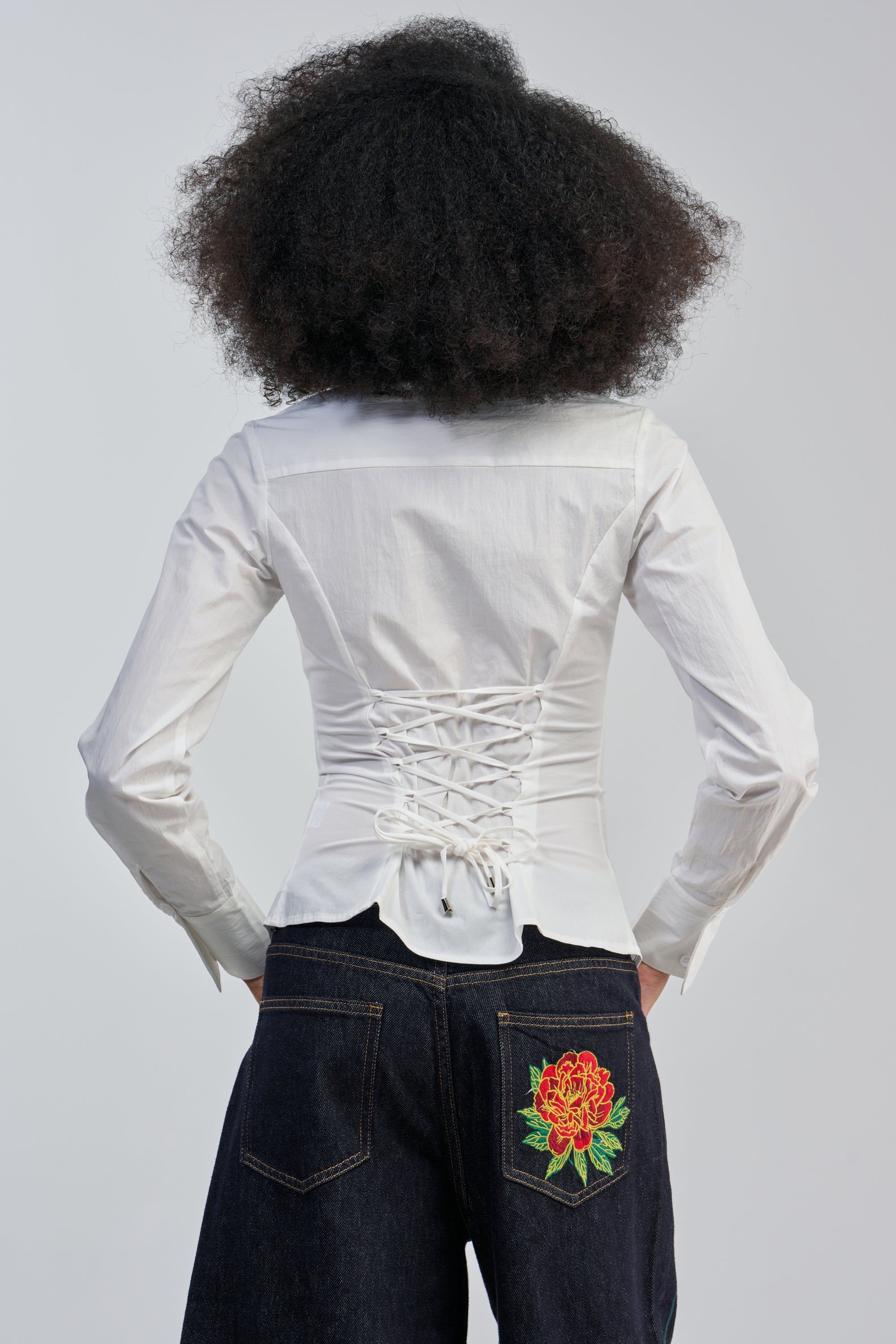 A person is seen from behind, wearing the OFFICE SIREN POPLIN BLOUSE—a white poplin blouse with a lace-up back—paired with dark jeans that have a large red flower embroidered on the back pocket, set against a light gray background.