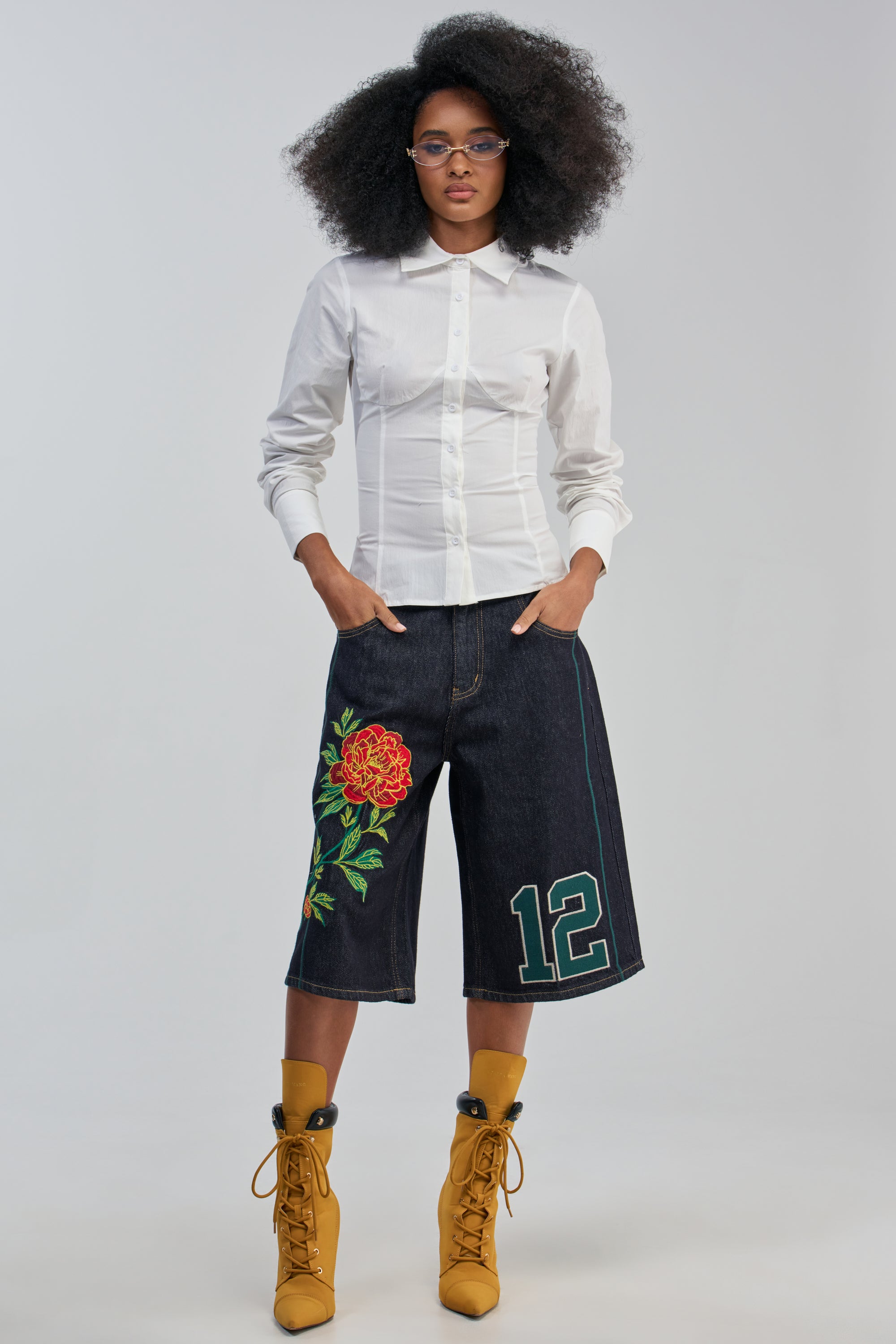 Woman with natural hair wears the sculpted fit OFFICE SIREN POPLIN BLOUSE in white, paired with wide dark denim shorts featuring a floral patch and number 12, yellow lace-up boots, and glasses, standing with hands in pockets against a plain background.