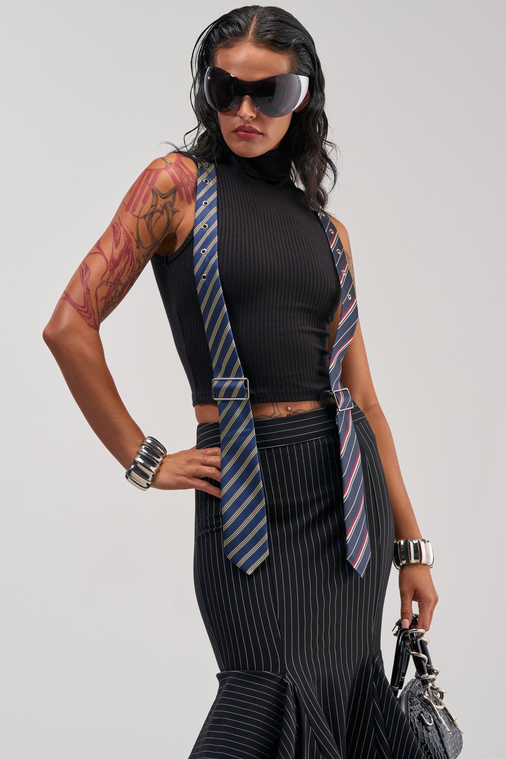 Wearing the TIED UP SLEEVELESS SWEATER—a black ribbed knit turtleneck—this individual styles it with large sunglasses, a pinstripe skirt, two striped neckties as suspenders, bold bracelets, and a textured handbag. The look is workwear-inspired and finished with a visible arm tattoo.