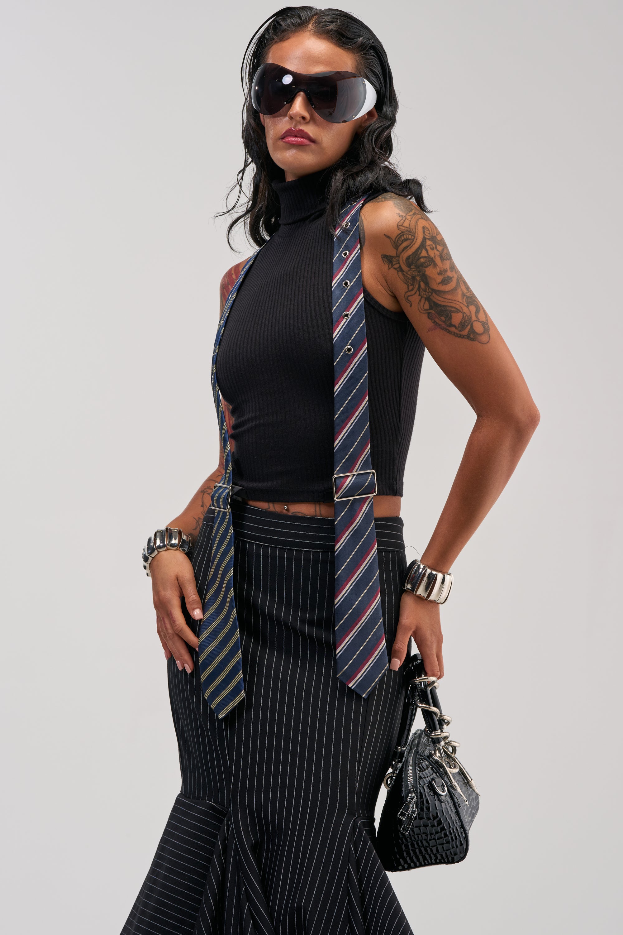 Wearing the TIED UP SLEEVELESS SWEATER, a woman in sunglasses with visible tattoos pairs it with two neckties and a pinstripe skirt, channeling workwear-inspired style as she confidently poses with a black purse.