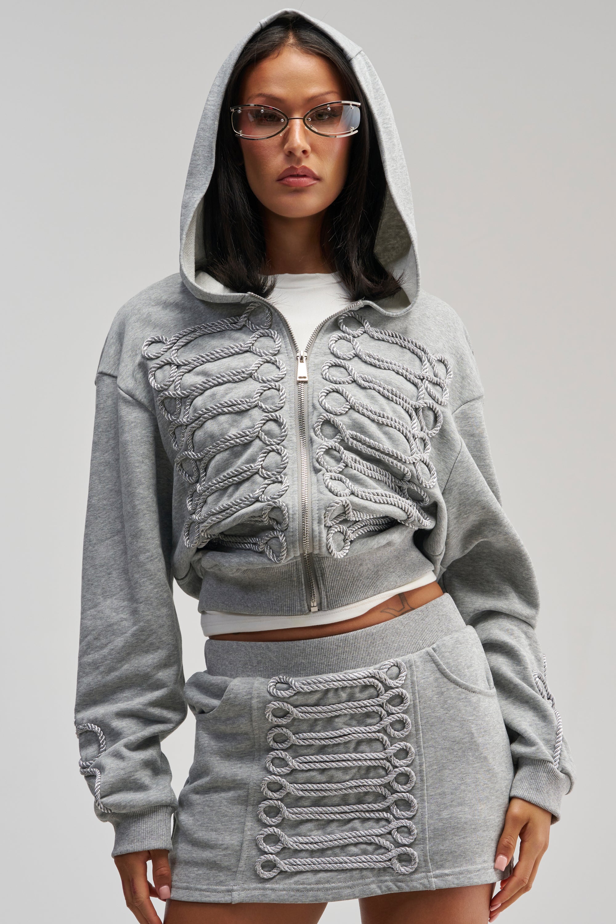 A person is wearing the JORDY FRENCH TERRY ZIP UP SWEATSHIRT in grey, styled with a matching gray skirt that features coordinating decorative details for a cohesive loungewear statement. They have straight dark hair, glasses, and a neutral expression.