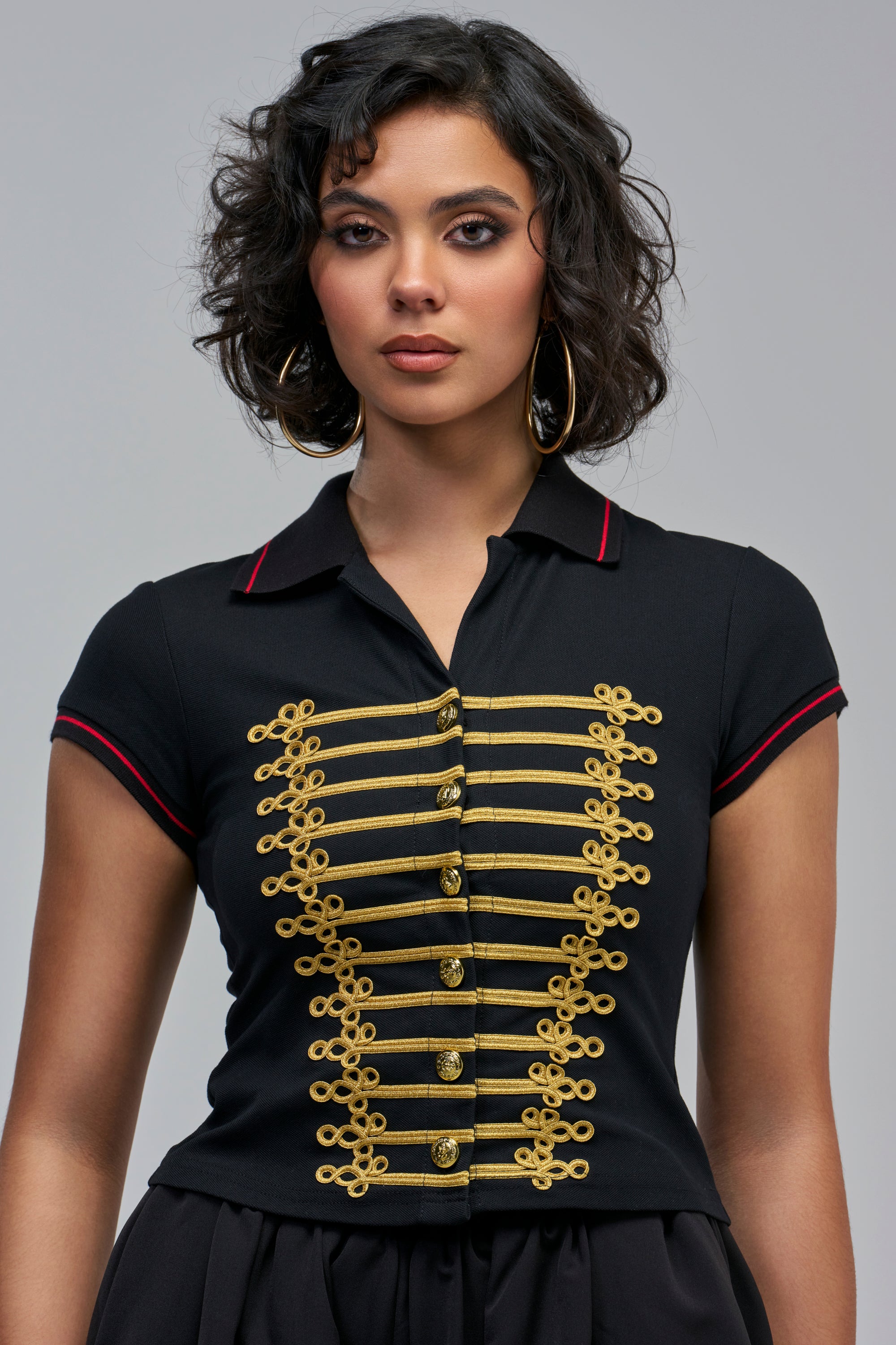 A woman wearing a black TOP SPOT KNIT POLO TOP featuring military-inspired gold braided trim and brass-colored buttons down the front.