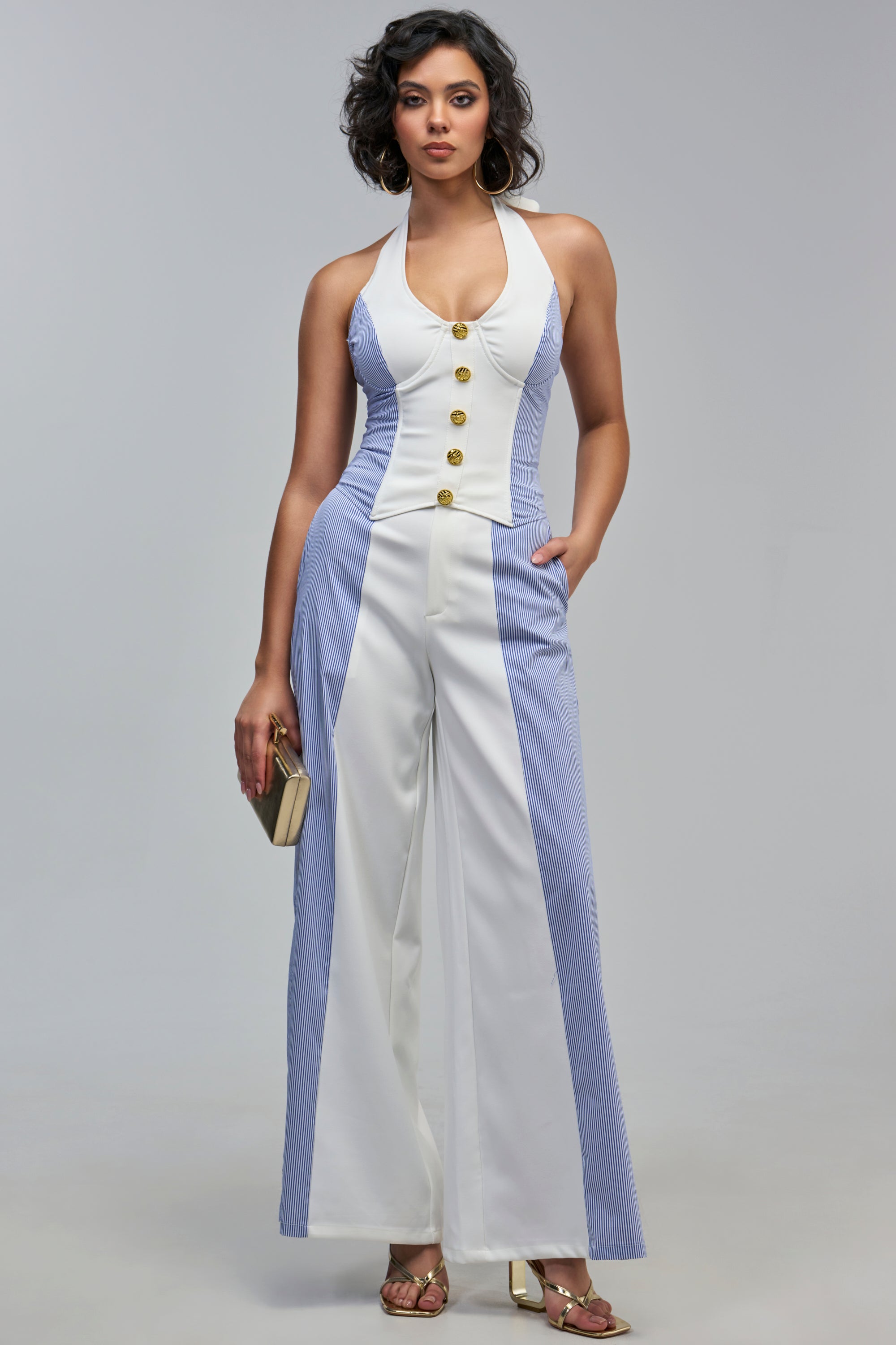 A woman wears THE SWEETEST GIRL RELAXED PANT as part of a halter-neck jumpsuit with a white bodice, gold buttons, and pinstripe wide-leg pants, while holding a small gold clutch.