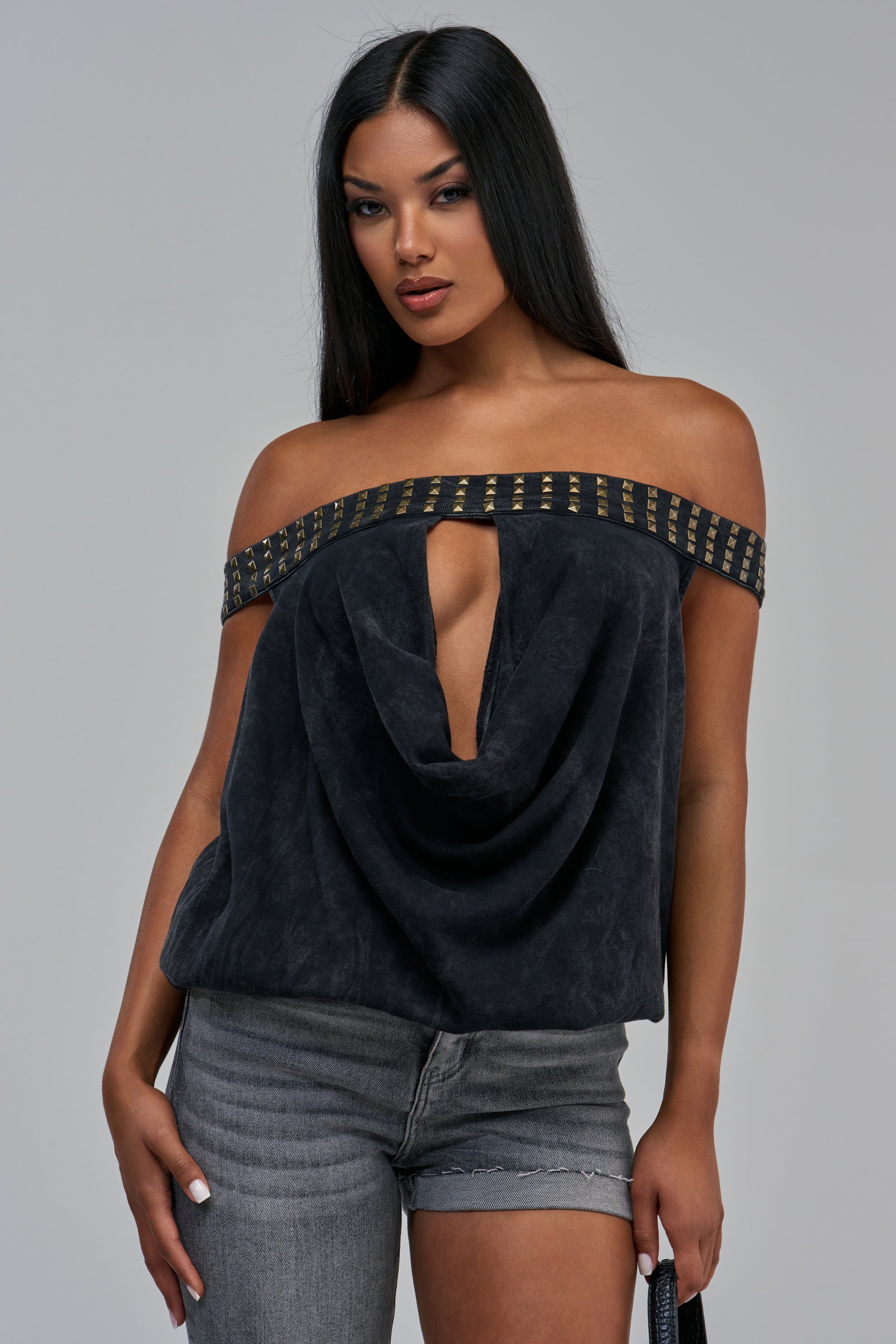 A woman wears the IN MY OWN LANE OFF THE SHOULDER SWEATSHIRT in washed black with a central cutout and metal-studded neckline band, paired with gray denim shorts.