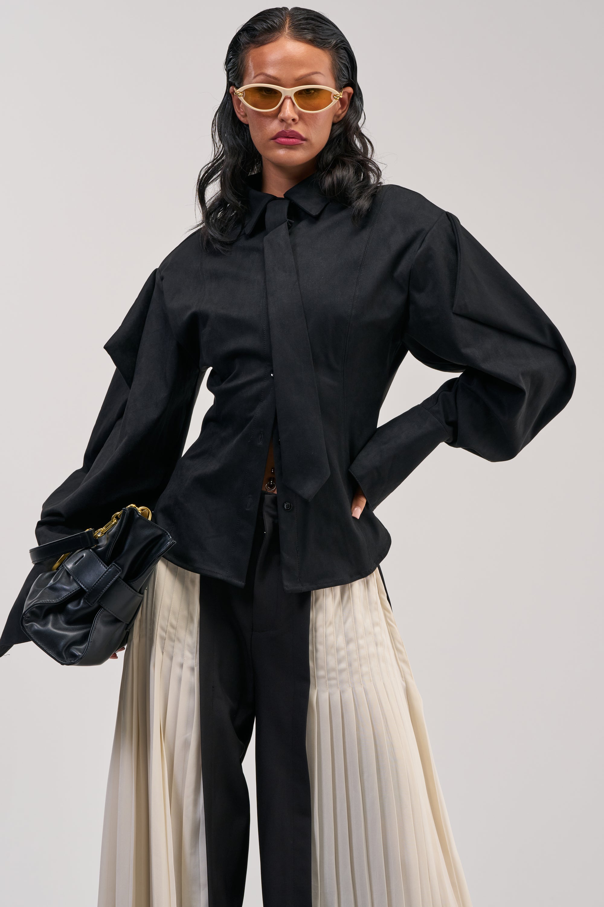 A woman wearing the SHE MEANS BUSINESS SUEDE BUTTON DOWN with oversized sleeves, black pants with white pleated panels, yellow sunglasses, and holding a black clutch stands against a plain background.